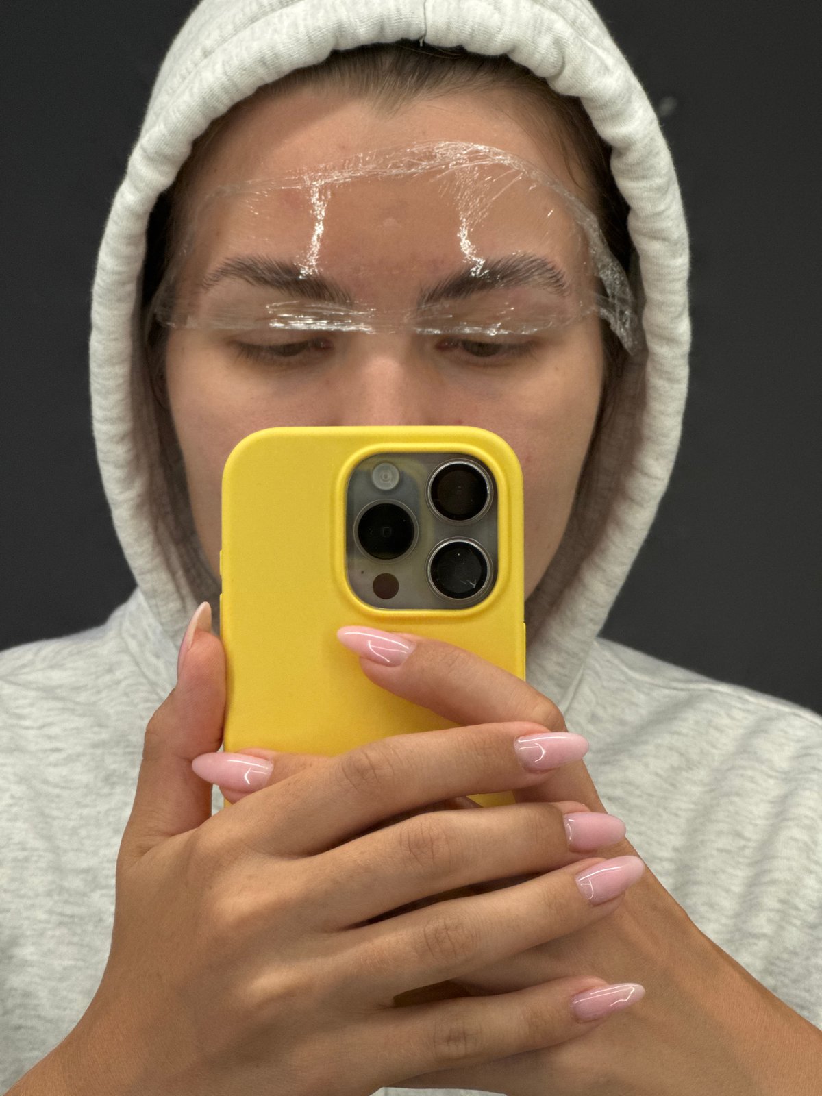 Person takes mirror selfie with yellow phone in gray hoodie