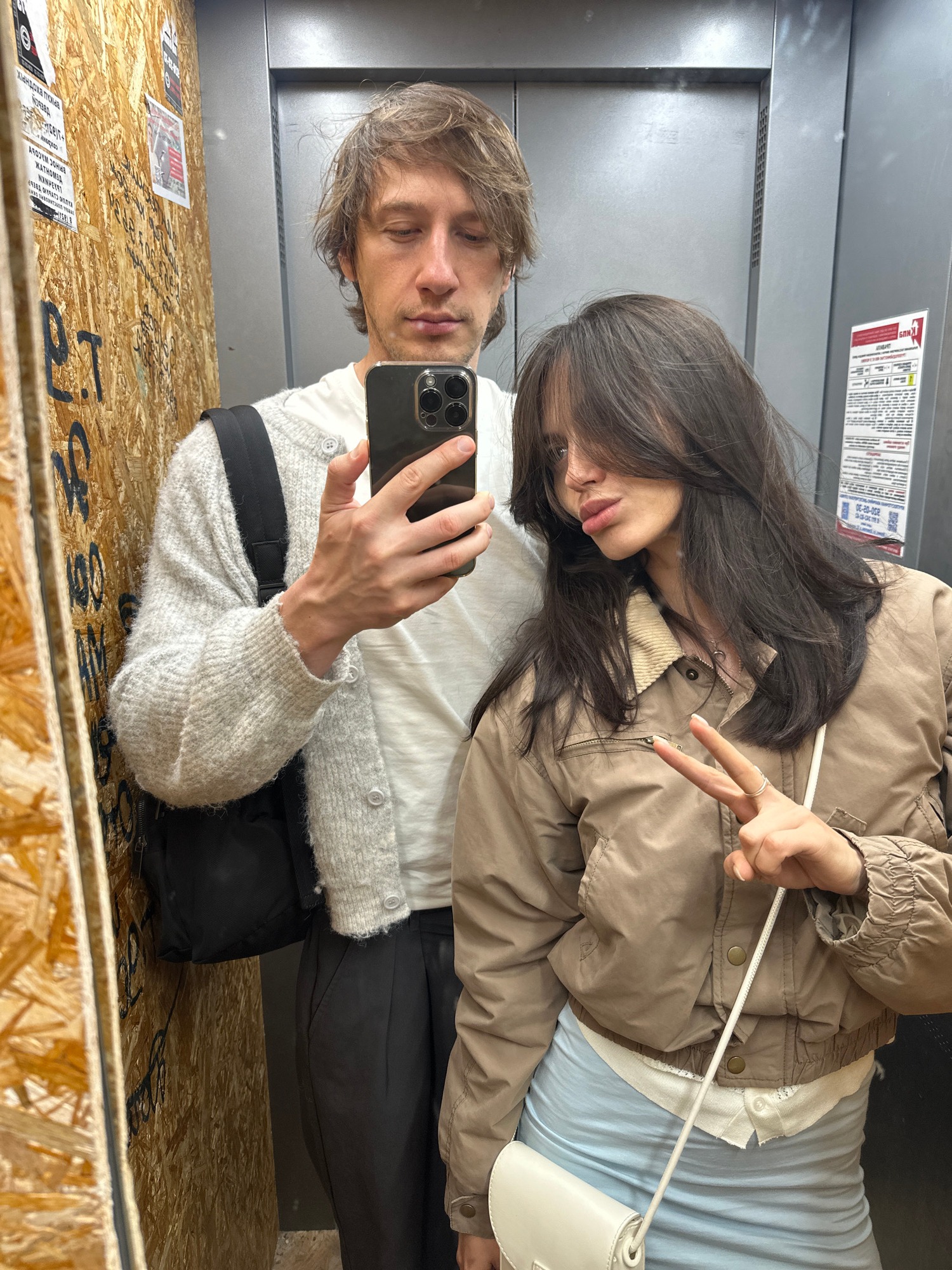Elevator mirror selfie, casual fits, smartphone in hand.