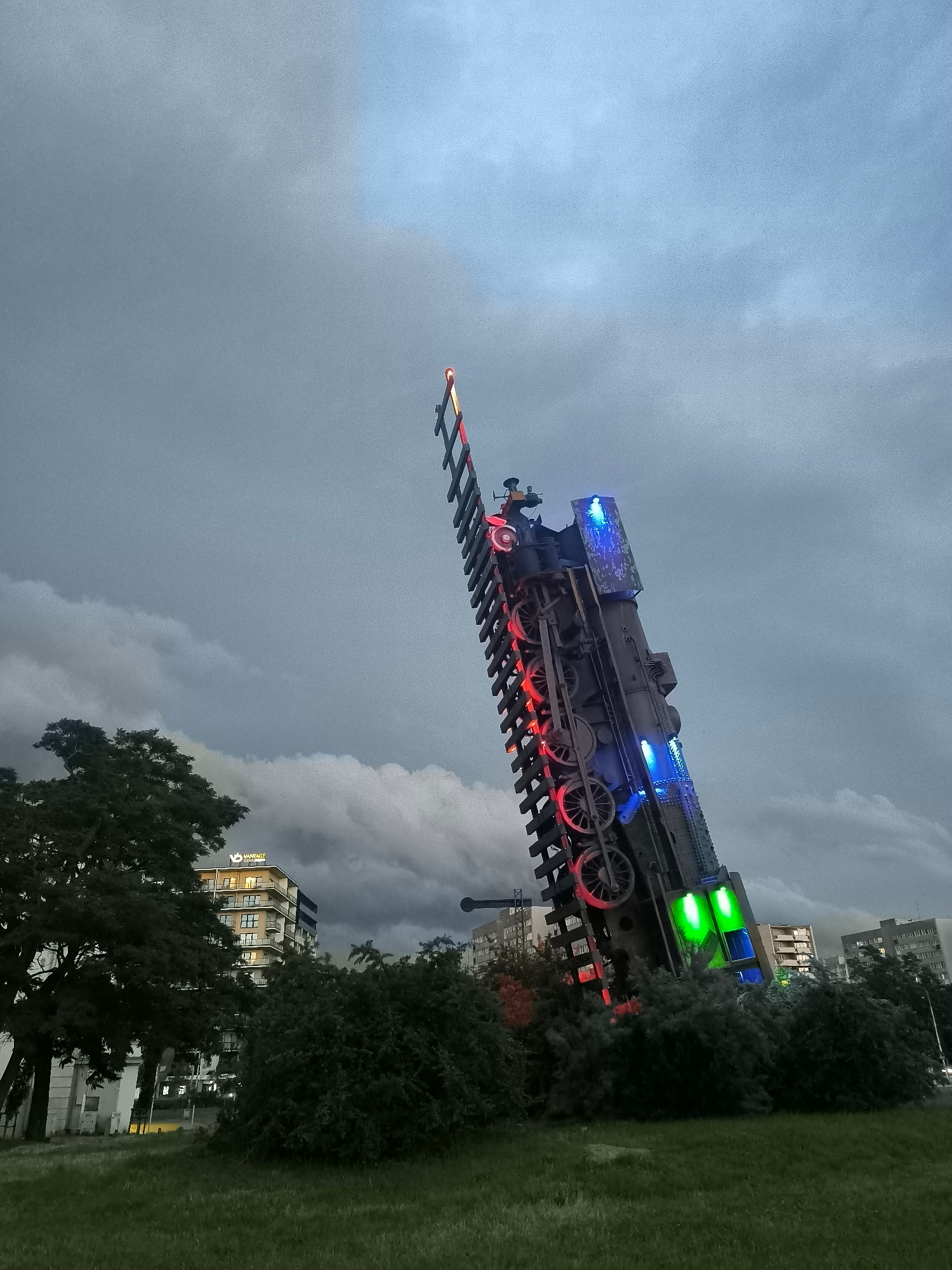An art installation resembling a train, tilted vertically with its front end pointing skyward, is prominently featured in the foreground of the image. This installation is illuminated with red, blue, and green lights. The scene is set against a cloudy, overcast sky. In the background, there are trees and buildings, including a multi-story building with the "Vantage" logo visible. The location is the Wrocław University of Economics and Business campus in Wrocław, Poland. The ground in the foreground is covered with grass. No people are visible in the image.