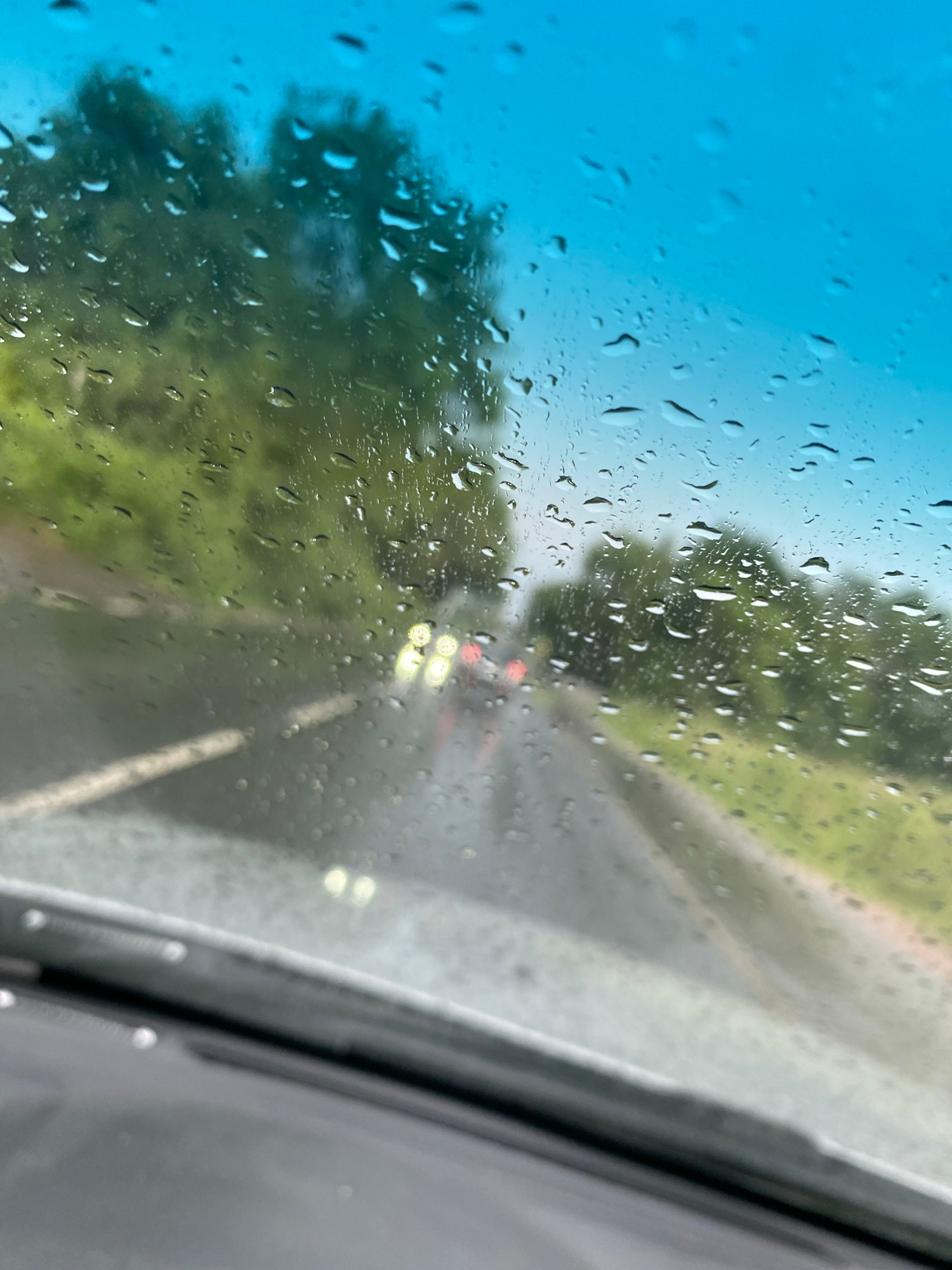 The image shows a view from inside a vehicle through a rain-streaked windshield. The sky is a vibrant blue, transitioning to a lighter shade towards the horizon. On the left side of the windshield, numerous raindrops are visible, distorting the view of the landscape beyond. To the right of the blue sky, a blurred green area suggests trees or foliage. Further to the right, a road is depicted, with a vehicle's headlights visible in the distance, suggesting it is either dusk or the weather is poor, causing reduced visibility. The road appears wet, consistent with the rainy conditions. The right edge of the frame shows the interior of the car, specifically a portion of the dashboard or window frame, which is out of focus. The overall impression is of a car journey on a rainy day. The location context is Przęsocin, Poland.