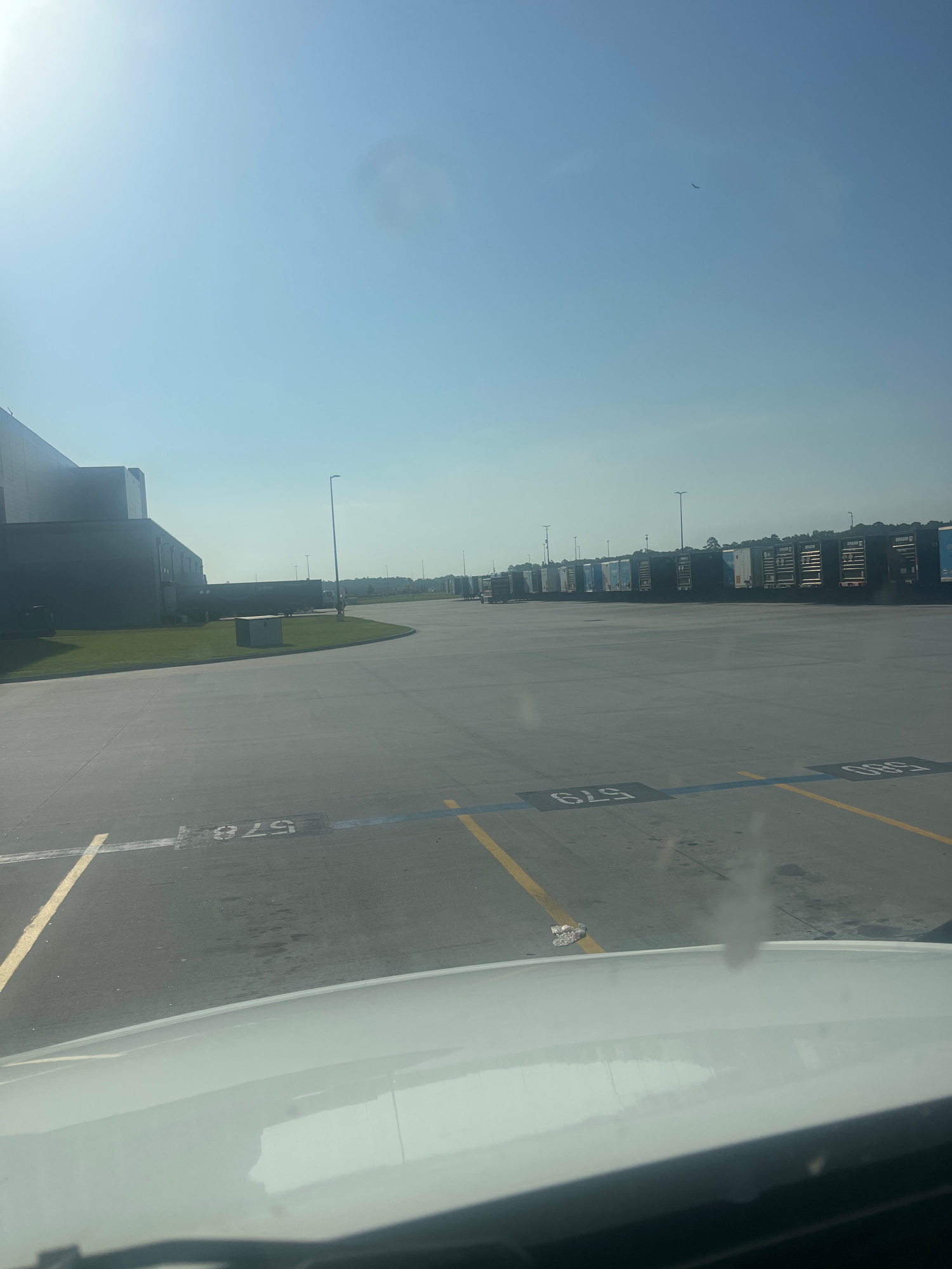 This is a low-angle, landscape-oriented photograph taken from an airplane window, showing an airport tarmac with various objects and the sky. The image is rotated 90 degrees clockwise.
The primary subjects are the tarmac, a line of parked semi-trailer trucks, and a vast, clear blue sky with a single bird in flight. The tarmac is marked with yellow and blue lines, and visible text includes the numbers "579" and "578" painted in black on the tarmac, likely indicating parking spots or taxiway identifiers. There are also some smaller buildings and structures visible in the distance, along with lampposts. A white airplane wing is prominently visible on the right side of the frame, indicating the perspective is from inside an aircraft. There are some patches of grass near the parked trucks. The lighting suggests it is daytime, possibly morning or afternoon, with bright sunlight casting a slight glare. The weather appears clear and sunny. The presence of the airplane wing and tarmac markings strongly suggests the location is an airport, specifically Pooler, United States of America. No people are clearly visible in the image. The overall mood is one of travel and observation, with a sense of spaciousness due to the open sky and tarmac.