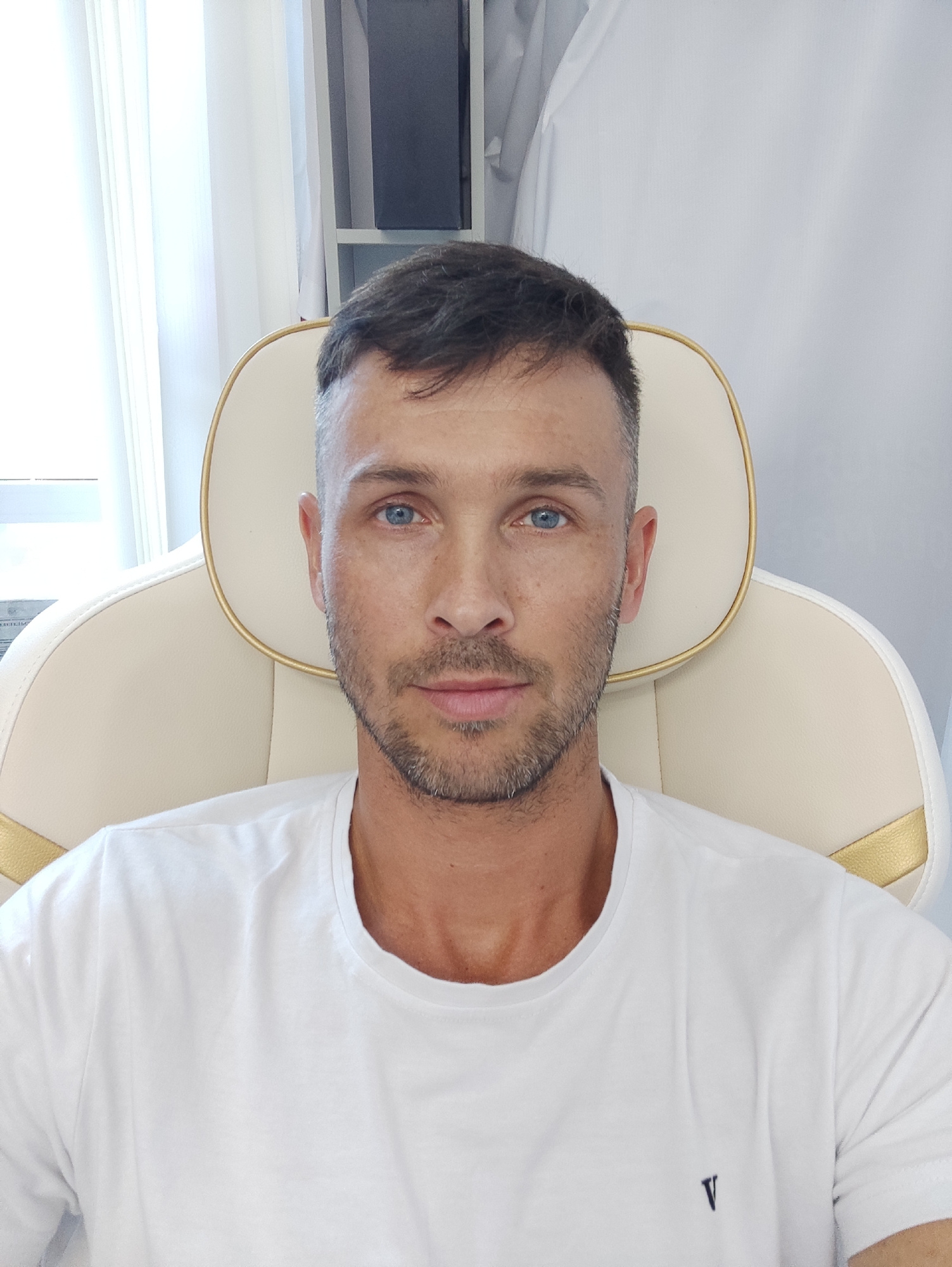 A medium shot shows a man with short, dark hair, blue eyes, and a light stubble beard sitting in a beige and gold upholstered chair. He is wearing a white t-shirt with a small, dark V logo on the left chest. The man is looking directly at the camera. The background is a white wall with sheer curtains to the left and a shelf with some objects to the right. The lighting appears to be natural, coming from the left.