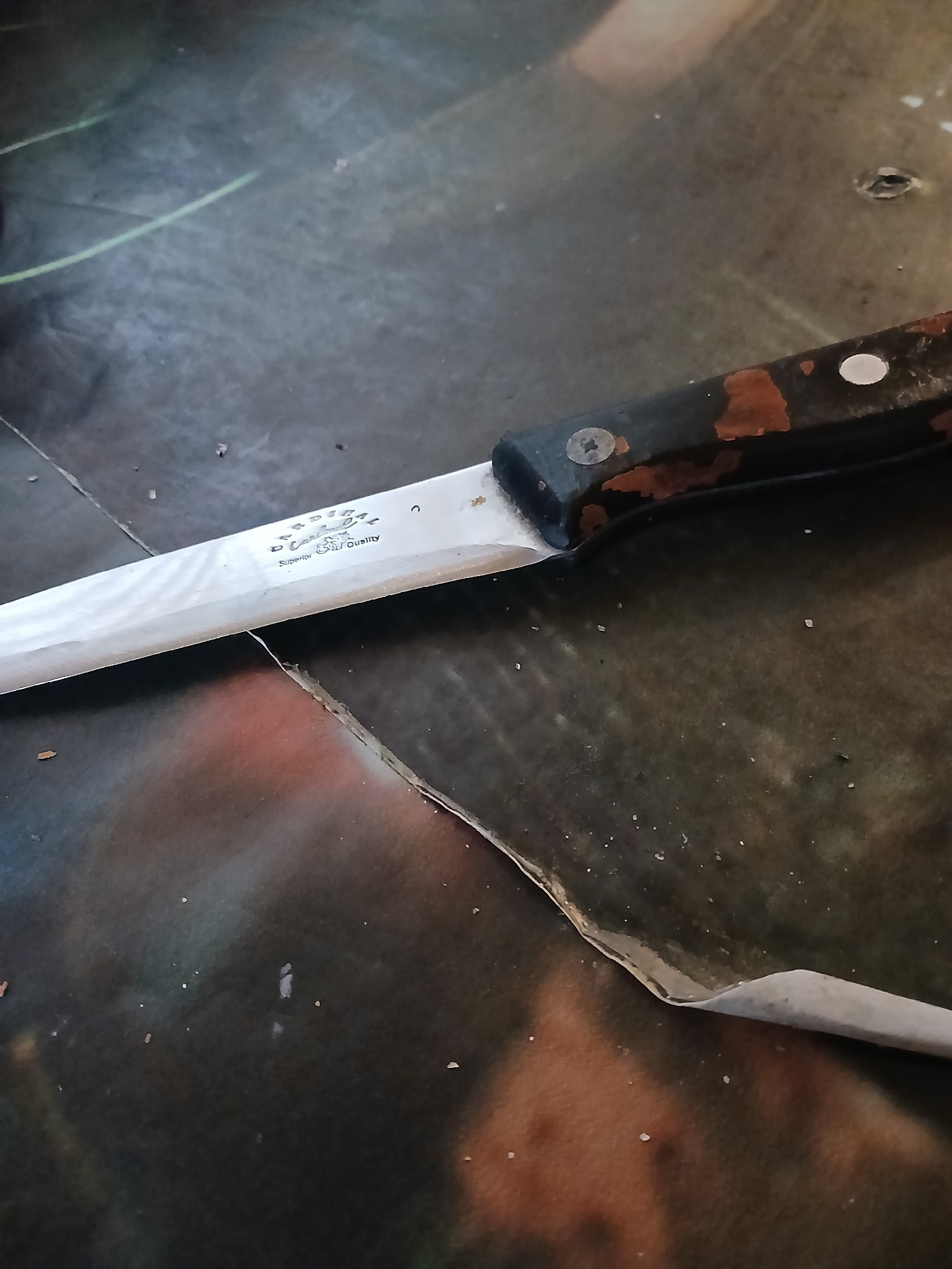 An old, somewhat rusted kitchen knife lies on a dark, patterned surface, possibly a table or a large piece of printed material. The knife has a black handle with visible wear and tear, and what appears to be some orange rust spots. The blade is metallic and appears to have some text or a logo etched into it, with the words "Superior Chef Quality" clearly visible. The surface it rests on is textured and has areas of lighter and darker shades, with a significant fold or tear visible in the lower right portion of the image, revealing a lighter colored material underneath. There are small, scattered white specks on the surface, which could be dust or debris. The lighting is dim, casting shadows and highlighting the textures of the knife and the surface. The overall impression is of an object in an indoor setting, likely domestic, given the nature of the item and its condition. There is no indication of the location being the city of Hlushky, Ukraine.