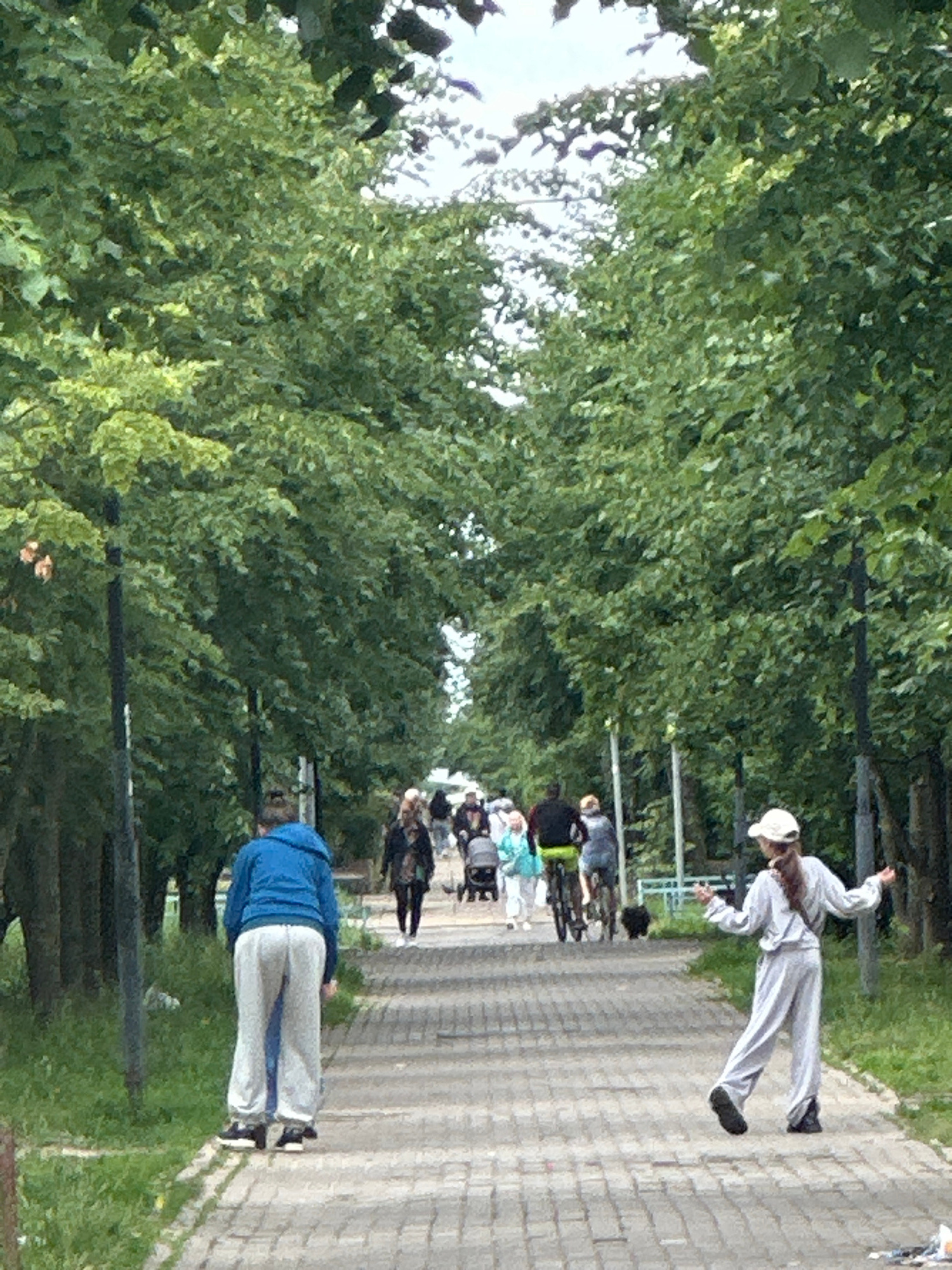 This is a photograph taken from a slightly elevated perspective, showing a group of people walking along a paved path in what appears to be a park or a tree-lined walkway. The setting is outdoors, with lush green trees and foliage dominating the left side of the frame, suggesting a warm-weather season, likely spring or summer. The path is made of light-colored paving stones and is bordered by green grass on both sides.
The main subjects are several individuals in motion. In the foreground, a person wearing a blue hooded jacket and light-colored pants is walking away from the camera. Further down the path, a group of people are walking in the same direction. One individual in the mid-ground is wearing a white outfit and a white hat, striking a dynamic pose as if mid-stride or mid-movement. Other people in the background are less distinct but appear to be a mix of adults and possibly children. The overall mood seems relaxed and casual, as if people are enjoying a walk or participating in an outdoor activity.
The time of day appears to be daytime, with bright lighting casting some shadows, though the presence of abundant green foliage suggests it might be overcast or the sun is not directly overhead. There are no discernible signs or text visible in the image. The location is identified as Podolsk, Russia, which, combined with the scenery, suggests a common urban or suburban park environment.