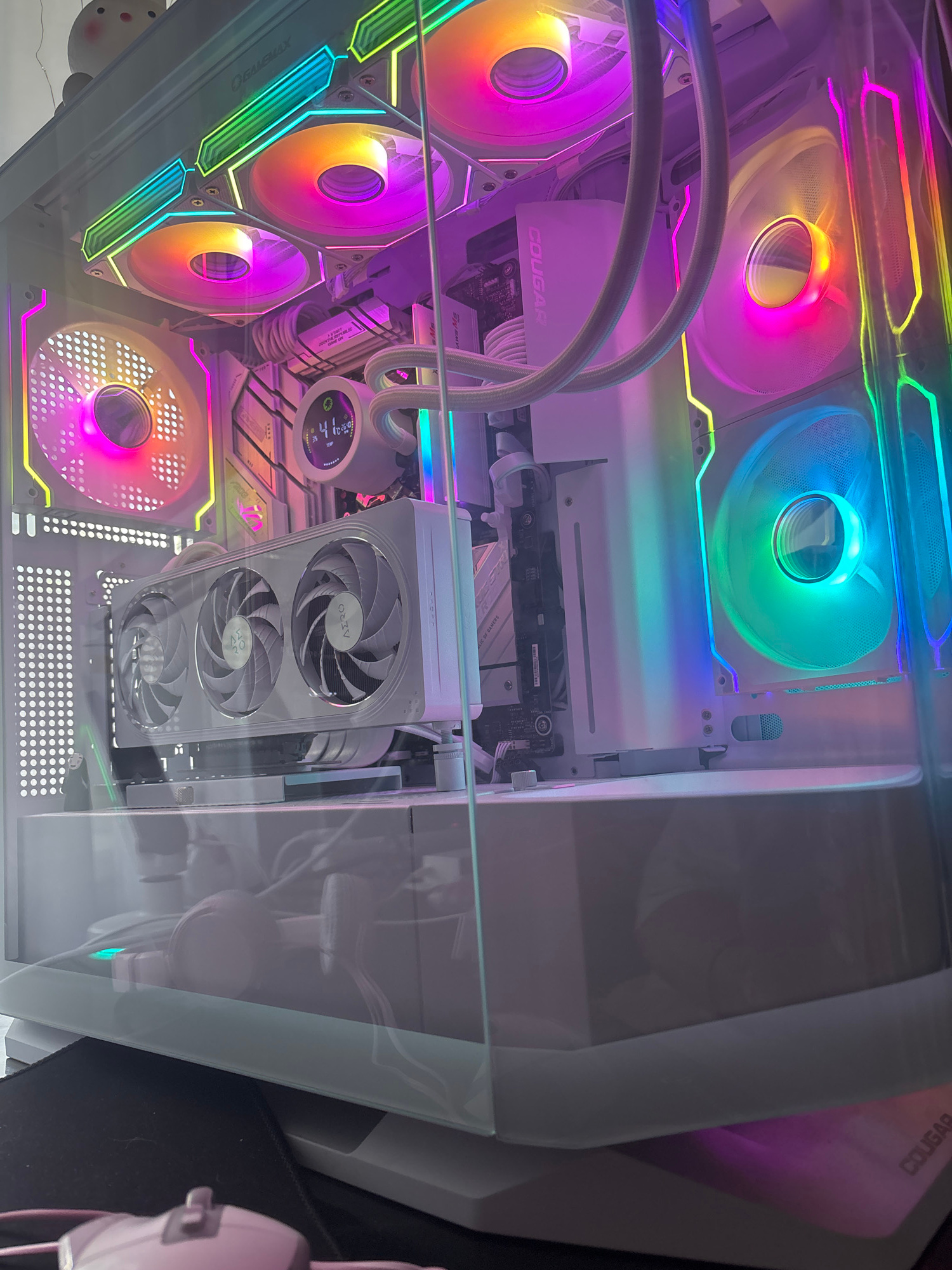 A white PC case with a clear side panel displays its internal components, illuminated by vibrant RGB lighting. The case contains a graphics card with three white fans, a CPU cooler with a circular digital display showing "41 10:53", and multiple case fans also featuring RGB lighting. The background shows a blurred, plush toy and the exterior of the PC case. The scene is indoors and appears to be a close-up shot of the computer.