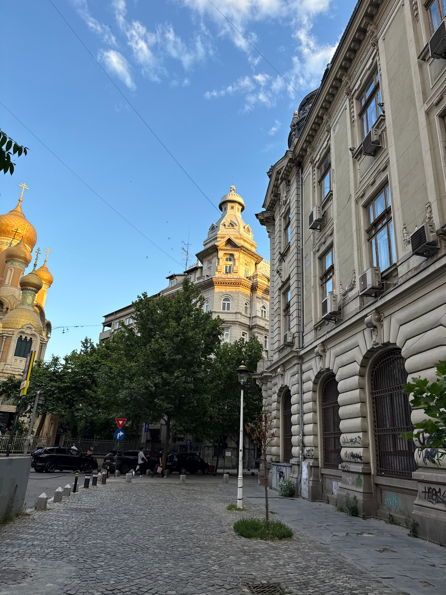 The image captures a street scene in Bucharest, Romania, featuring architectural elements of both religious and secular nature. On the left, the golden domes and crosses of a Romanian Orthodox church are prominently visible, bathed in the warm glow of the late afternoon sun. Adjacent to the church, a block of apartments stands, its facades relatively plain. To the right, a grand, ornate building with classical architectural details lines the street, its windows featuring intricate carvings and iron grates. Several air conditioning units are mounted on its facade. The street itself is paved with cobblestones, and a narrow sidewalk runs along the right side of the image. Parked cars, mostly black SUVs, are lined up along the curb, and a few pedestrians are walking on the sidewalk. Traffic signs, including a yield sign and a directional arrow, are visible at an intersection. The sky is a clear blue with a few wispy clouds, and some trees with lush green foliage add to the urban landscape. The overall impression is that of a city street with a blend of historical and modern elements, under a bright, clear sky.