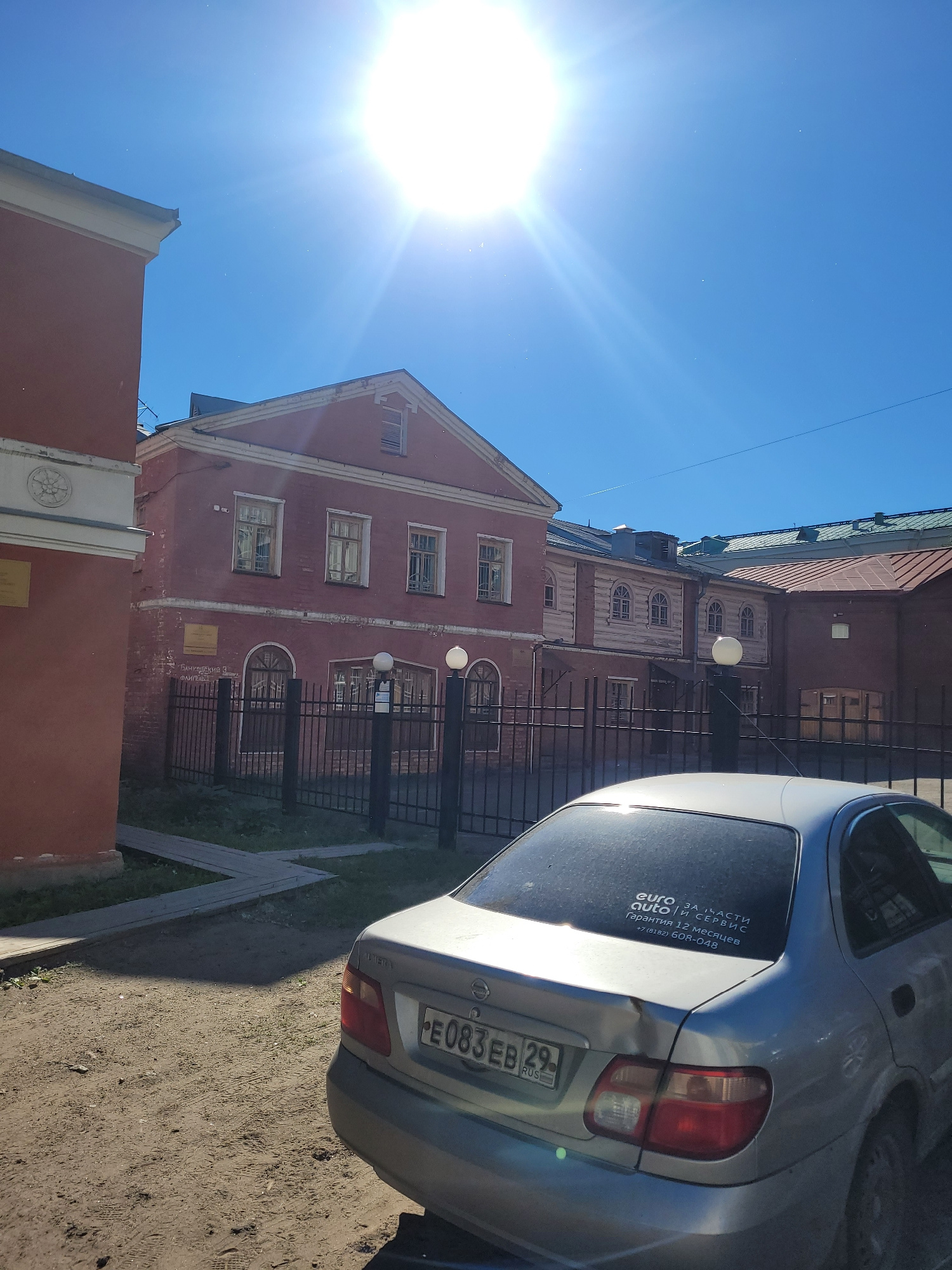 The image shows a sunny day in Arkhangelsk, Russia, with a clear blue sky. In the foreground, a silver sedan is parked on a dirt road. The car has a license plate that reads "E083EB 29 RUS" and a sticker on the rear window that says "euro auto запчасти и сервис гарантия 12 месяцев +7 (8182) 608-048".
Behind the car, a low black metal fence separates the street from a pathway and a grassy area. Beyond the fence, there are several old brick and wooden buildings, characteristic of historical architecture. One building is painted red and has multiple windows. Another section of the building appears to be made of light-colored wood with arched windows. The buildings have pitched roofs. The sun is very bright and is positioned high in the sky, creating lens flare and casting shadows. The overall impression is that of an urban environment with older structures, bathed in strong sunlight.