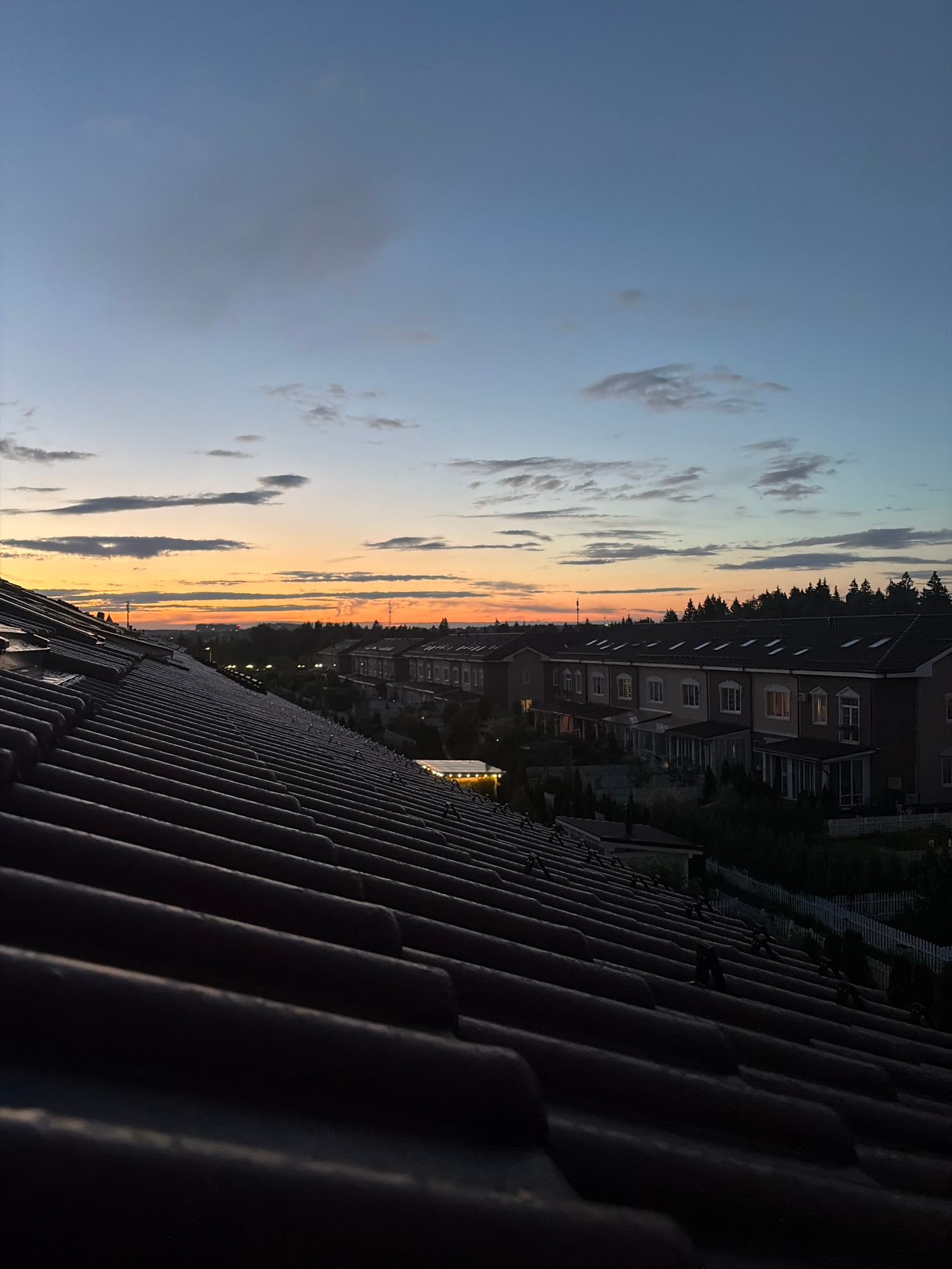 An elevated perspective captures a twilight scene over a residential area in Baranovo, Russia. The foreground is dominated by a dark, tiled roof, angled diagonally to the right, its texture and rows of tiles clearly defined in the dim light. Beyond the roof, a line of terraced houses stretches across the middle ground. These buildings are predominantly light-colored with dark roofs and evenly spaced windows, some of which are illuminated from within. A white picket fence runs along the front of the properties. Behind the houses, a dense line of dark green trees forms a backdrop, suggesting a wooded area. The sky above is a gradient of soft blues and grays, with streaks of orange and yellow near the horizon, indicating the setting sun. Scattered clouds, ranging from wispy to more substantial, drift across the sky. The overall impression is one of a quiet, suburban evening. There are no people or animals visible in the image.