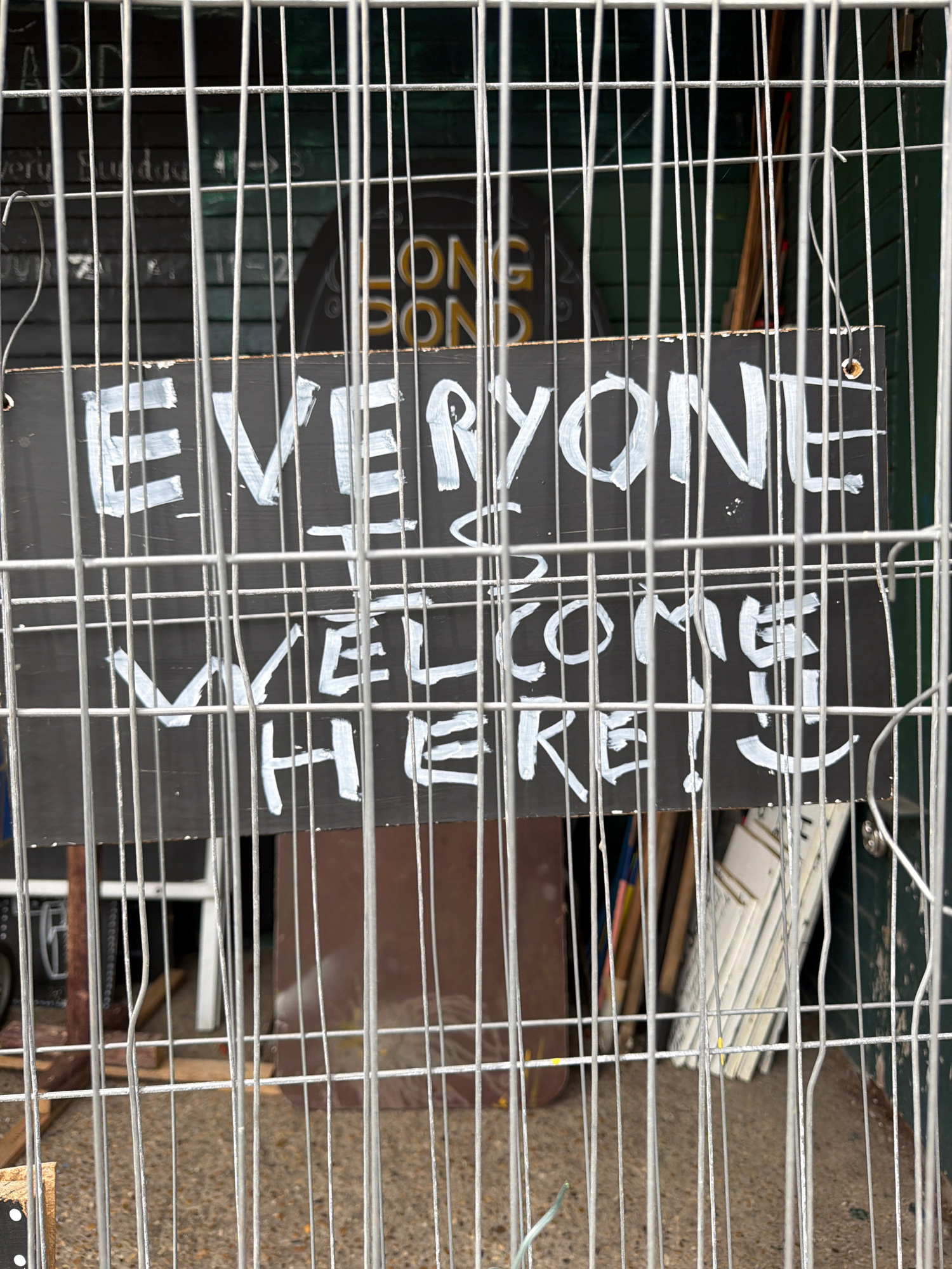The image shows a black sign with white text partially obscured by a metal grate. The sign appears to be outdoors, possibly in a storage area or construction site, with a concrete ground visible in the background. There are also some stacked items and wooden planks visible behind the grate. The lighting suggests it is daytime.
The text on the sign is written in a somewhat messy, hand-painted style. It reads: "EVERYONE WELCOME TO ENJOY THE COFFEE." Below this, there's another line that is harder to decipher, but appears to be "NON STOP FUN".
The overall impression is a casual, perhaps informal, welcome message on a sign that is not centrally displayed or highly polished. The grate in front of it suggests the sign might be part of a temporary setup or in a location that requires some form of barrier.