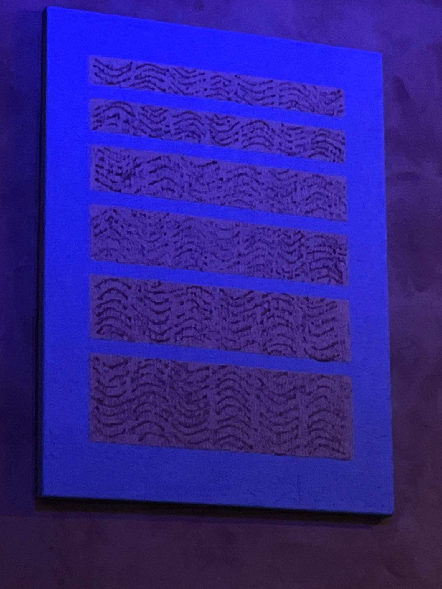An abstract painting with a bright blue background features six horizontal rectangular sections filled with a textured, dark purple pattern. The pattern in each section consists of repeating, wavy, parallel lines that create a somewhat organic or naturalistic appearance, possibly mimicking wood grain or ripples. The painting is displayed on a wall, which appears to be a dark, textured surface. The overall lighting suggests a dimly lit environment, possibly with a blueish hue casting onto the scene. The painting's location in Mũi Né, Vietnam is not evident from the visual information provided.