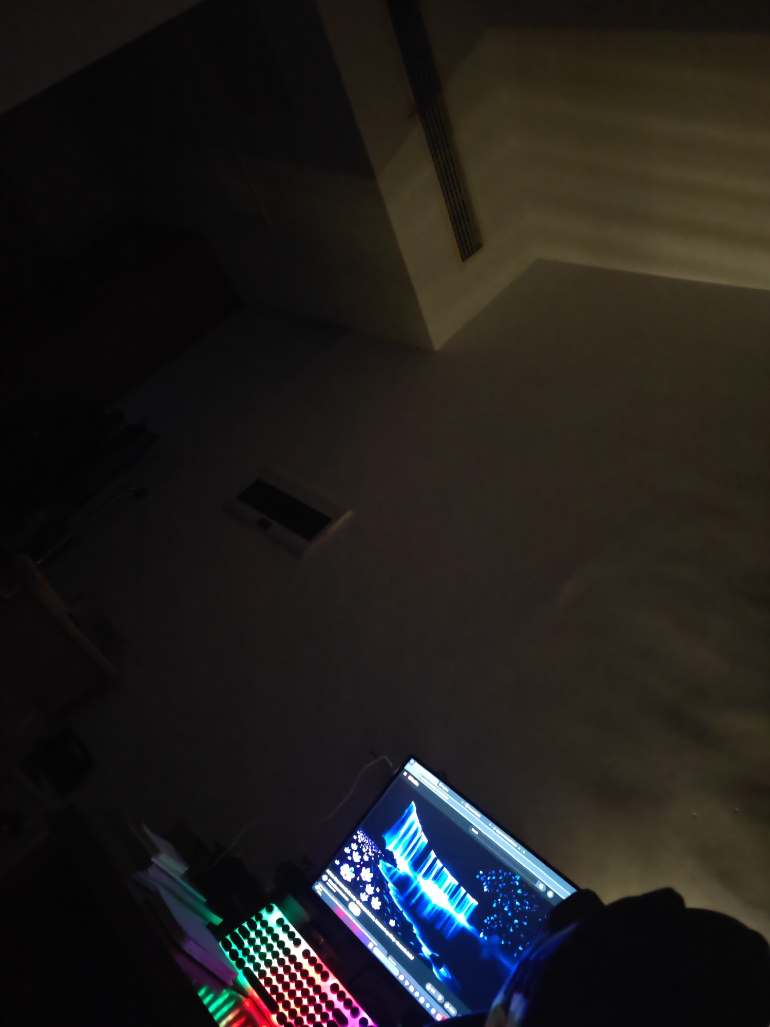 A dimly lit room in Jiangning District, China, features a computer setup with a monitor displaying a vibrant blue and white abstract image. In the foreground, a mechanical keyboard with colorful RGB backlighting is visible, suggesting a gaming or professional setup. The overall ambiance is dark, with the computer's illumination providing the primary light source. The ceiling shows a ventilation grate, and the walls are bare.