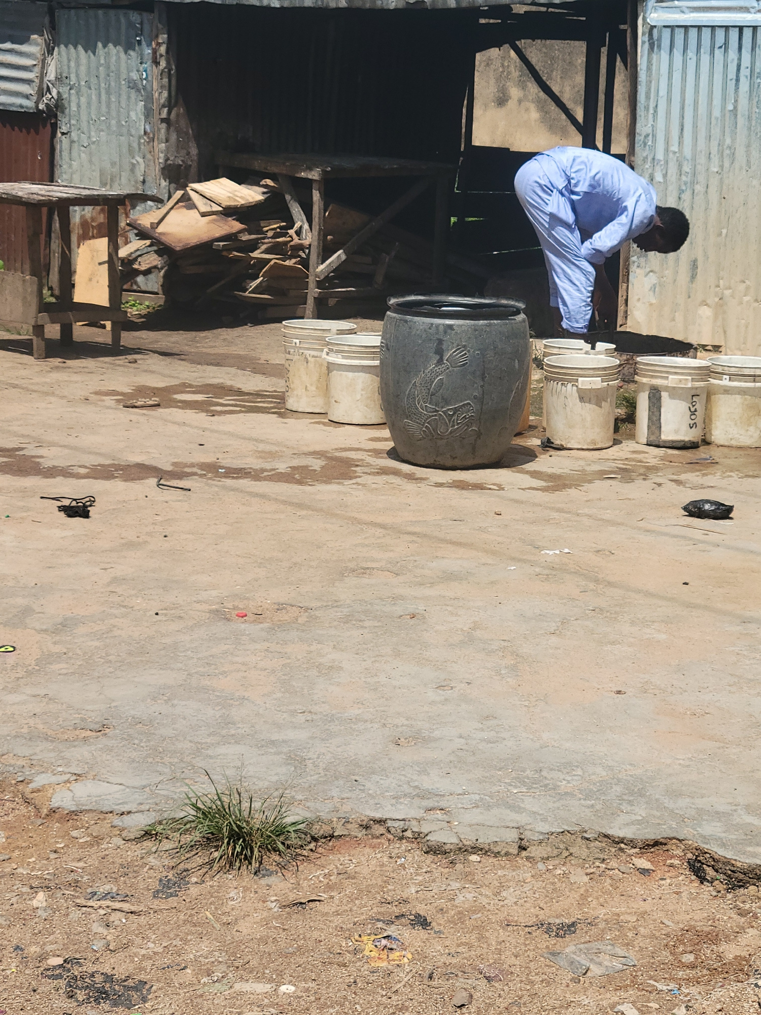 In the city of Jos, Nigeria, a man dressed in a light blue garment is bending over in front of a large, dark gray cylindrical container. The container has a carved image of a fish on its side. Behind the man and the container are several white buckets, some with visible labels, and a pile of wooden planks. The scene appears to be outdoors, with a weathered, corrugated metal structure in the background and a concrete ground surface that shows signs of wear and dirt. There are also scattered debris and a patch of dry grass in the foreground.