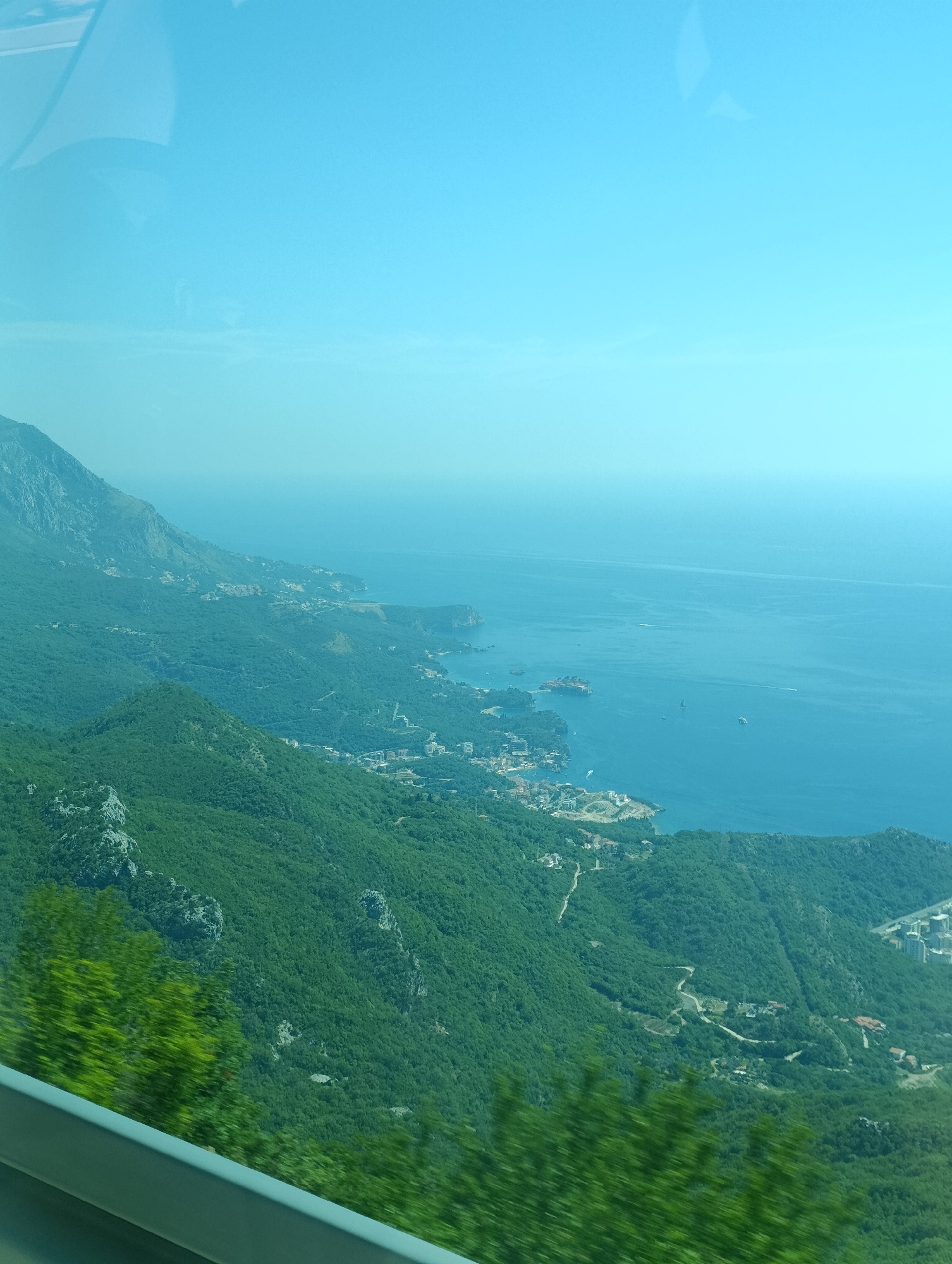 An aerial, wide-angle, slightly hazy view captures the coastal landscape of Brajici, Montenegro. The scene is dominated by lush green, densely forested mountains that cascade down to meet the bright blue Adriatic Sea. A serpentine road winds its way through the hills, with scattered buildings and structures visible amidst the trees and along the coastline. The sea itself is calm, with a few small boats dotting the surface and a distant, hazy horizon line where the sea meets the pale blue sky. The image appears to have been taken from a moving vehicle, possibly a bus or car, due to the blurred foliage in the foreground and the reflection of what looks like a window frame in the upper left corner.