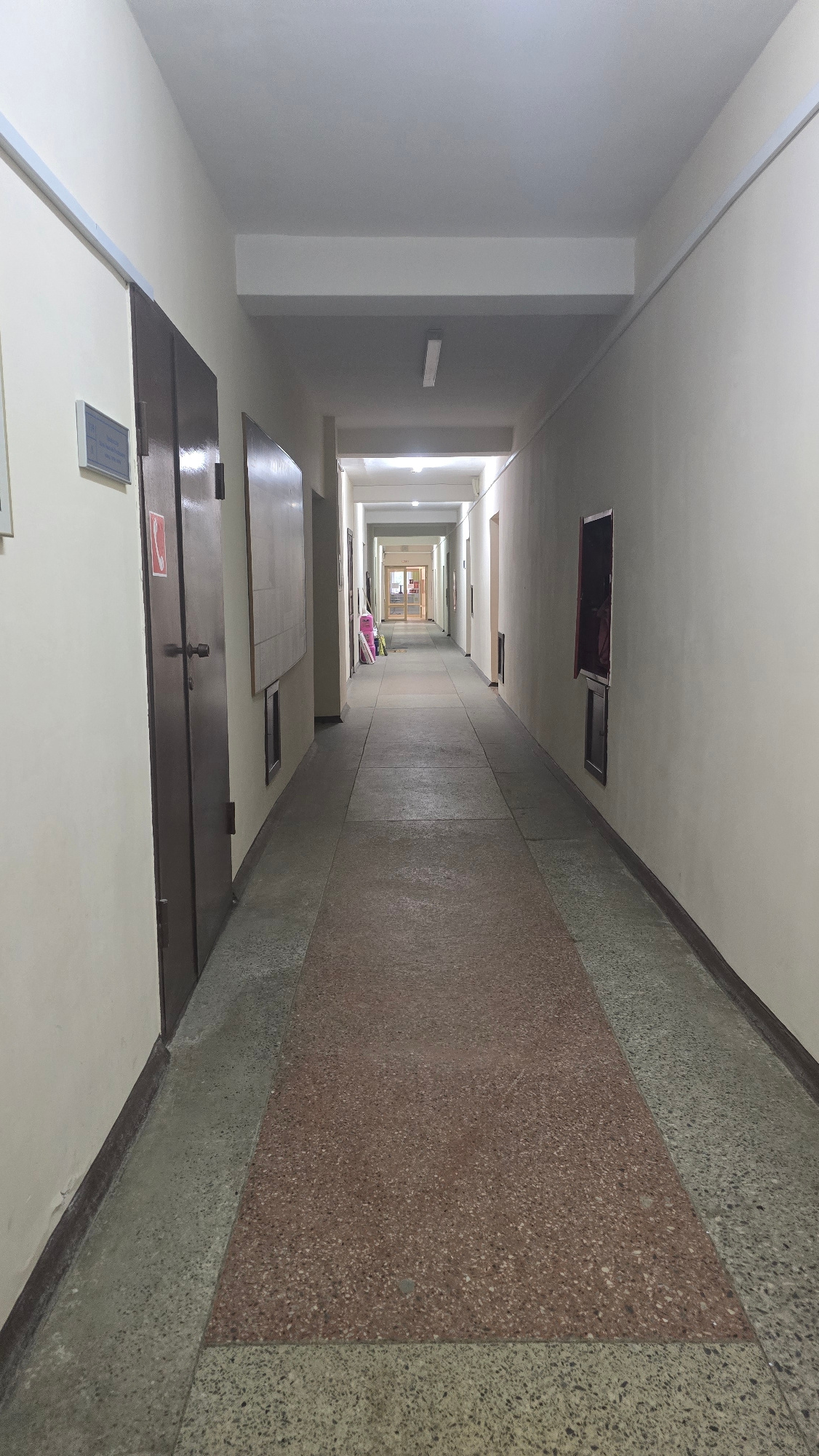 This media file shows a long, brightly lit corridor looking down from a high angle, giving a sense of extreme perspective. The corridor appears to be in a building with multiple floors, as suggested by the receding openings in the ceiling and the repeating pattern of doors and walls. The walls are a neutral color, and the floor is covered in a speckled terrazzo material, with a large rectangular inlay of reddish-brown terrazzo in the center.
The corridor is sparsely furnished. In the foreground, there are several dark brown doors, one of which has a red and white fire exit sign visible. There are also some flat, dark surfaces that could be tables or shelves. Further down the corridor, near the center, are a few items that appear to be stacked luggage or bags, in shades of pink and white. At the far end of the corridor, a set of glass doors leads to another section of the building, which is also illuminated.
The overall impression is one of institutional architecture, possibly a dormitory, apartment building, or hospital. The lighting is bright and even, suggesting it is daytime. There is no visible weather, as the scene is entirely indoors. The extreme perspective, with the corridor narrowing to a single point in the distance, creates a dramatic visual effect. The scene is devoid of people, contributing to a sense of stillness and perhaps slight emptiness.
Some text is visible: a red and white fire exit sign on a door in the foreground. Another small, rectangular sign with white text on a blue background is visible on a white surface in the foreground.