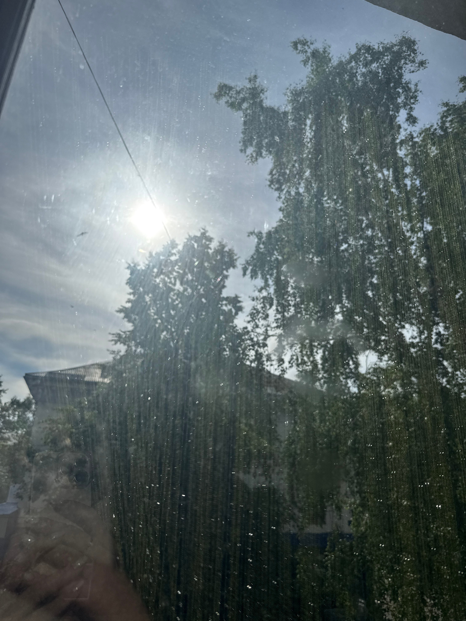 The image captures a sunlit sky with clouds viewed through a window, likely in Salavat, Russia. Tall trees with dense green foliage dominate the right side of the frame, their leaves appearing blurred and streaked due to reflections or imperfections on the glass. In the background, a building with a dark, sloped roof is visible, partially obscured by the trees. The sun is a bright, hazy disc in the upper left quadrant, its rays scattering across the sky. A thin wire stretches diagonally from the top left corner towards the sun. A reflection of a hand holding a smartphone with a camera array is present in the lower left corner of the image. The glass surface exhibits numerous vertical streaks and small, bright spots, suggesting it is dirty or wet.