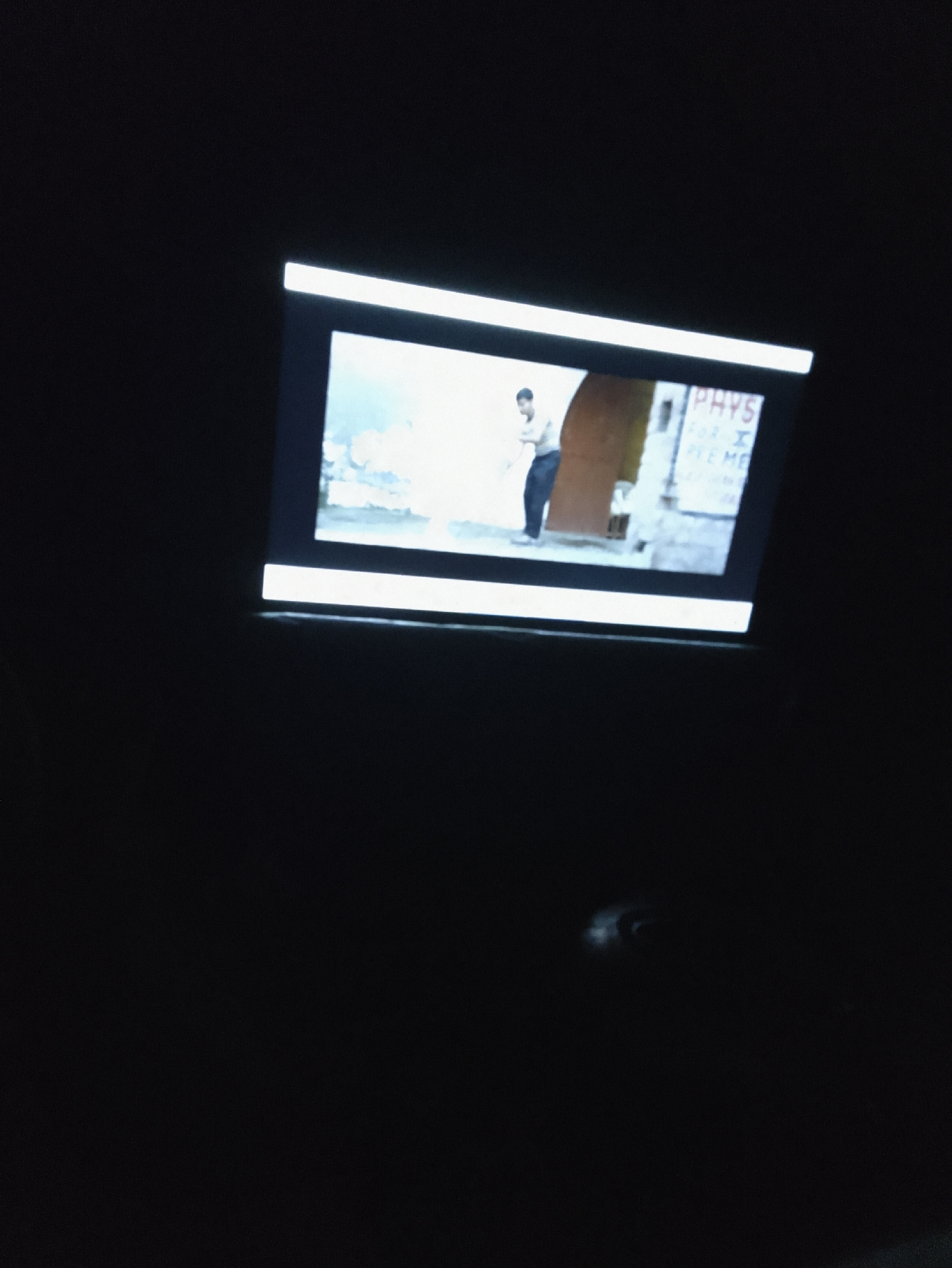 The image shows a close-up of a small, illuminated screen displaying a scene. The screen is framed by a bright white light at the top and bottom edges, suggesting it might be a projection or a screen within a device. The displayed scene appears to be outdoors or in a very bright, open space with a somewhat dilapidated appearance.
The main subject on the screen is a person, seen from the waist up, standing and facing slightly to the left. They are wearing a light-colored shirt and dark pants. To the right of the person, there is a vertical element that appears to be a doorway or a pillar, colored in a rough, textured brown. Behind the person and the doorway, the background is a bright, textured surface, possibly a wall, with some lighter patches that could be paint or light reflections.
On the right side of the screen, partially obscured by the doorway, is a sign with visible text. The text is in a blocky, capitalized font. The visible parts are: "PHYS" at the top, followed by "FOR I" on the next line, and then "PIE ME" on the line below. The letters are slightly blurred and difficult to read definitively. The overall scene has a somewhat grainy or pixelated quality, typical of older or low-resolution digital media. The surrounding area of the original image is very dark, indicating the screen is the primary light source and the photo was likely taken in a dimly lit environment.