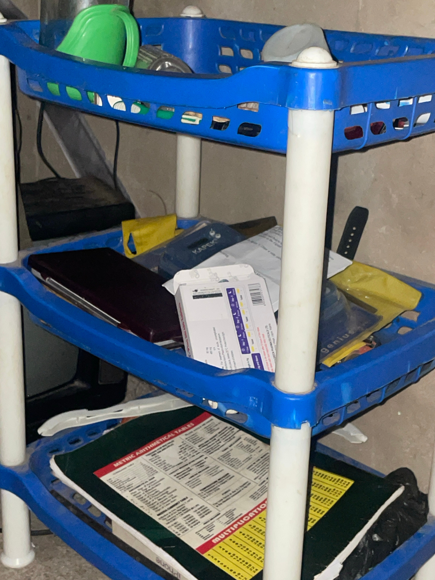 The image displays a blue, three-tiered plastic shelving unit containing various items. The top shelf holds a green cup and other indistinct objects. The middle shelf contains a dark red rectangular item, possibly a book or a case, a yellow packet, and some white packaging with text. The bottom shelf has a green item with red and white text that appears to be educational material, possibly a "Metric Arithmetical Tables" chart, and other items that are not clearly discernible, including a black plastic bag. The shelving unit is placed against a plain, light-colored wall, and the overall lighting suggests an indoor environment. The scene is static and shows no people or ongoing activities. The location is identified as Jalingo, Nigeria.