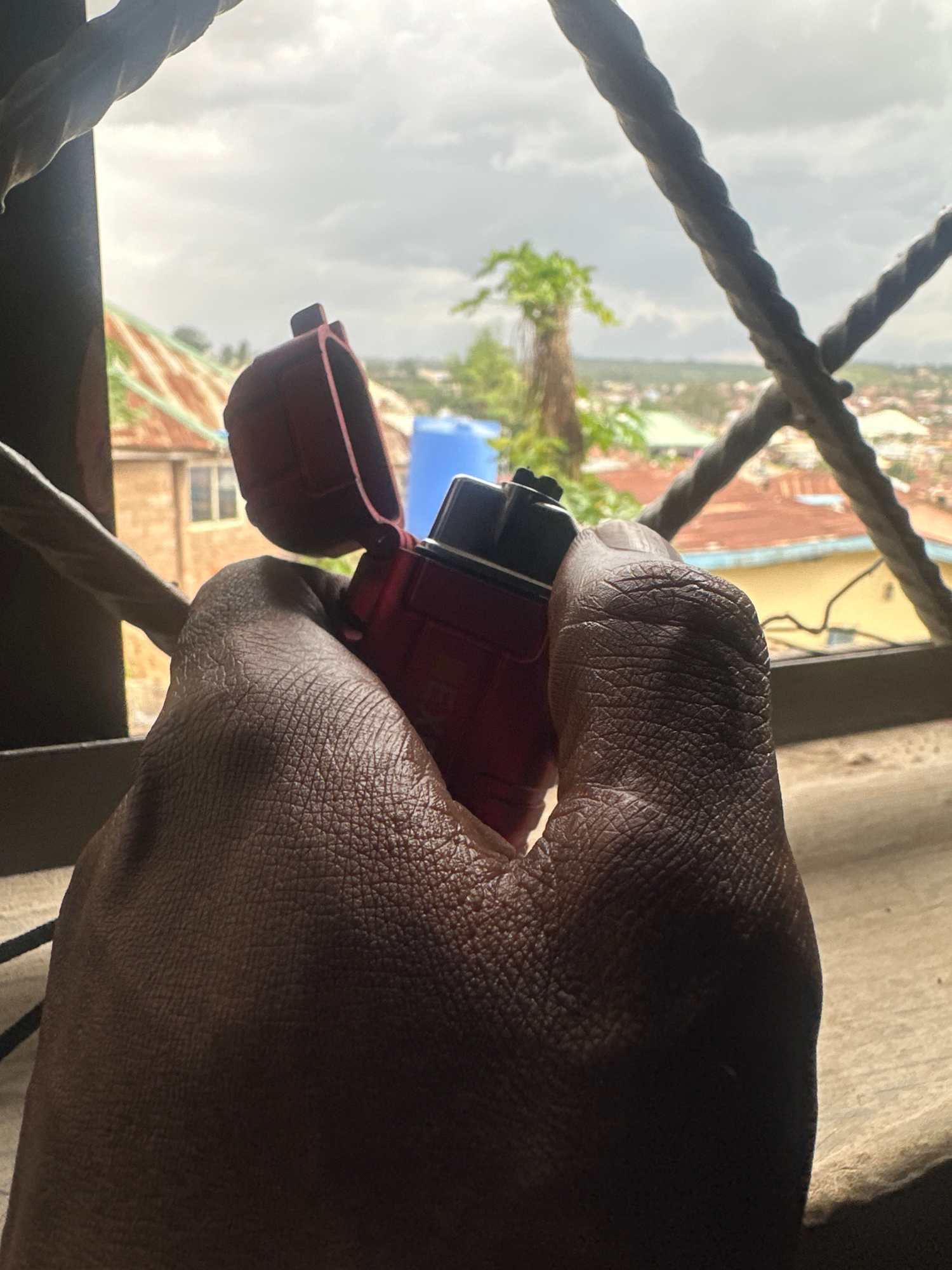 A close-up, low-angle shot captures a dark-skinned hand holding a red lighter with its lid open. The hand's texture is detailed, showing wrinkles and skin pores. The lighter is positioned in the foreground, partially obscuring a view of the background.
Beyond the hand and lighter, a window frame with two thick, dark metal bars divides the scene horizontally. Through the window, a blurred cityscape is visible under a cloudy sky. The city appears to be in Jos, Nigeria, with buildings featuring red-tiled roofs and some yellow walls. A bright blue water tank stands out amongst the rooftops, and a tall, thin tree with sparse green foliage is visible to the left of the frame. The overall impression is one of an indoor perspective looking out onto an urban environment.