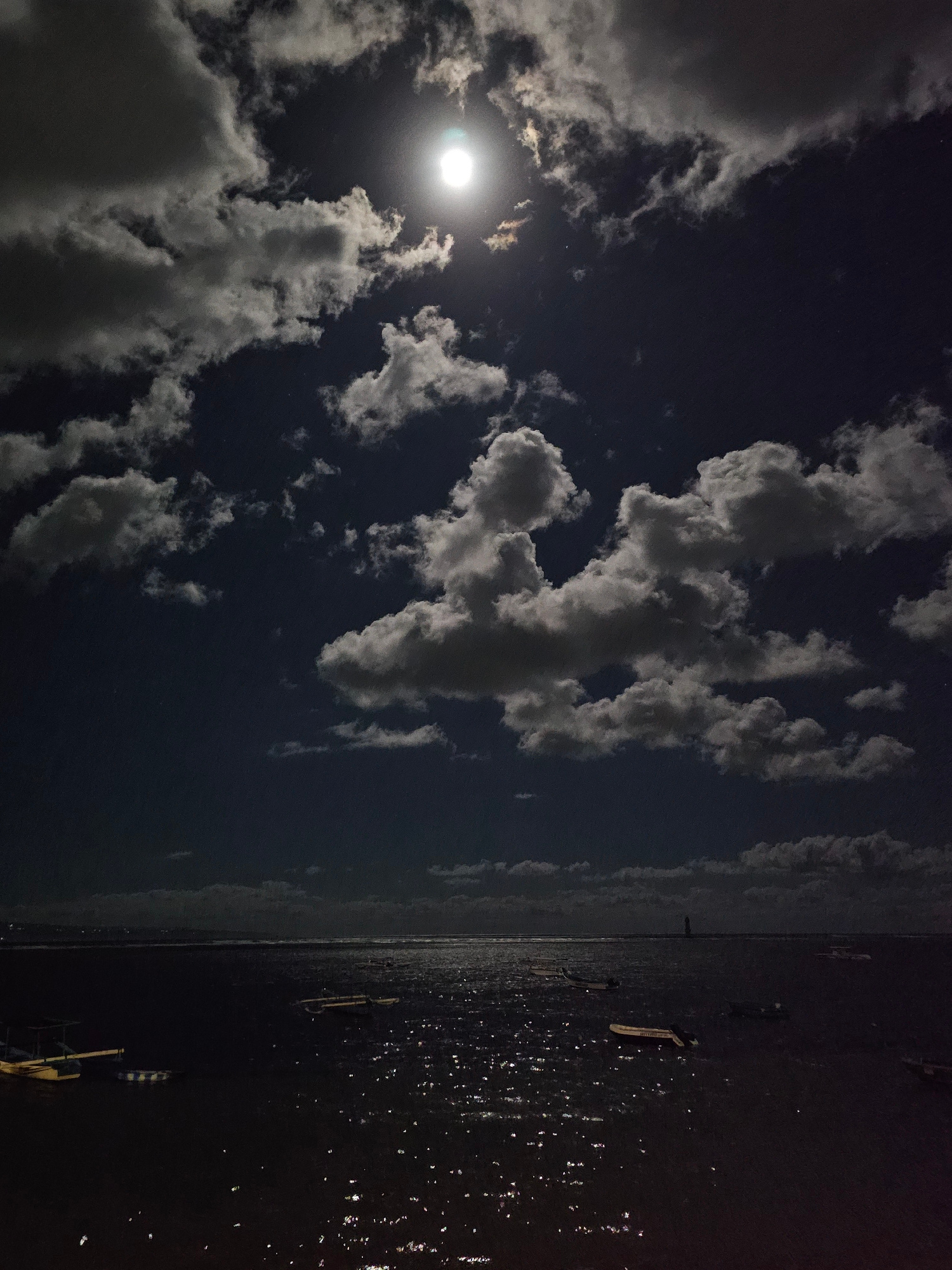 This media file captures a nighttime scene at sea near Denpasar, Indonesia. The image is oriented vertically with the sky on the left and the water on the right.
In the sky portion, a bright full moon is visible, partially obscured by scattered clouds. The clouds are illuminated by the moonlight, creating a dramatic contrast against the dark night sky.
The water area is dark and reflects the faint light from the moon and possibly distant lights from the shore. Several small boats are visible on the water, some appearing to be moored or anchored. The surface of the water is choppy, with many small points of light reflecting on it, suggesting ripples or waves catching the available light.
The overall mood is serene and somewhat mysterious, typical of a moonlit night by the ocean. The scene appears to be a still moment, with no apparent human activity. The location cues, such as the boats and the general seascape, are consistent with a coastal setting in Indonesia. The time of day is clearly night, indicated by the darkness and the presence of the moon. The weather appears clear enough for the moon to be visible, though clouds are present.