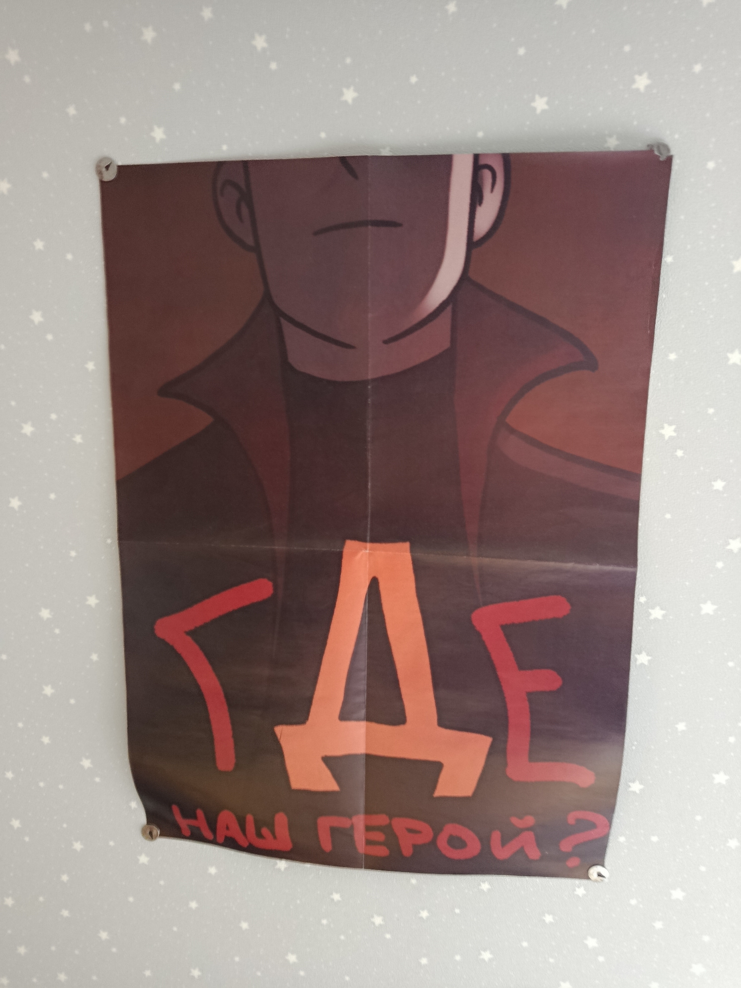 A poster is affixed to a wall with star-shaped patterns using metal thumbtacks. The poster features a stylized illustration of a person's upper body, with their face obscured by shadow, wearing a dark jacket with a prominent collar. Large, bold lettering in red and orange fills the center of the poster, spelling out the word "ГДЕ" (Gde), which translates to "WHERE" in English. Below this, in smaller red lettering, is the phrase "НАШ ГЕРОЙ?" (Nash Geroy?), meaning "OUR HERO?". The poster appears to be creased in the middle, suggesting it has been folded. The surrounding wall is light blue with a repeating pattern of small white stars. There is no discernible information within the image to indicate its location in Vitsebsk, Belarus.
