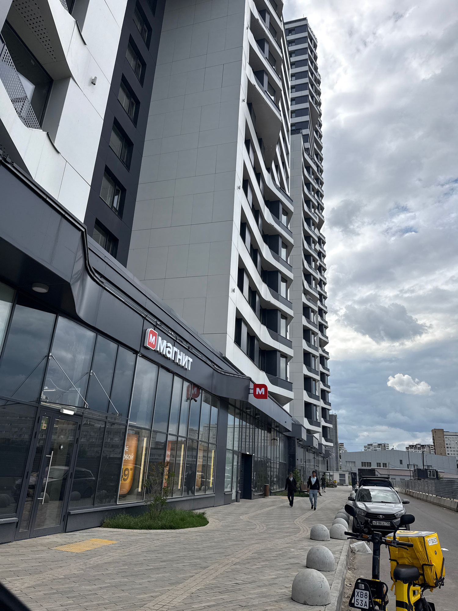 This image captures a contemporary building facade with a distinctive architectural style, characterized by its angular, layered design and a combination of dark and light gray panels. The building appears to be a residential or commercial complex, with rows of windows visible.
In the foreground, a black-clad building with large glass windows and a prominent "M Магнит" sign indicates a retail establishment. This sign is in a red square with a white "M" and white Cyrillic lettering. Reflections of buildings and the sky are visible in the glass.
Outside the building, a paved sidewalk runs alongside the facade, lined with rounded concrete bollards. A dark-colored car is parked near the sidewalk, and behind it is a bright yellow delivery scooter or motorcycle with a large insulated cargo box on the back, suggesting a delivery service is active. A license plate is partially visible on the scooter.
Several people are walking on the sidewalk. One individual in a light-colored jacket and dark pants is walking away from the camera, while another person in dark clothing is walking towards the camera. The weather appears overcast, with a cloudy sky dominating the upper portion of the image. The scene suggests a daytime setting in an urban environment. The overall atmosphere is modern and functional.
Visible text: "M Магнит", "H371BE 799" (car license plate), "XB8 53A" (scooter license plate), and some Cyrillic text on advertisements visible through the shop windows. The context provided indicates the location is Moscow, Russia.