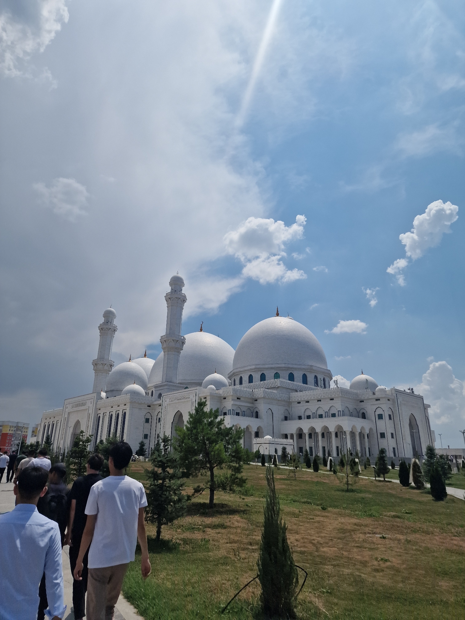 A wide-angle shot captures a large, white mosque with multiple domes and minarets under a bright, partly cloudy sky. In the foreground, several individuals are walking on a paved path towards the mosque. The mosque's architecture is intricate, featuring arched windows and decorative elements. The surrounding environment includes manicured lawns and scattered evergreen trees. The sky is a mix of blue and white clouds, with a lens flare visible in the upper left quadrant. This is the Seyitmeket Mosque in Shymkent, Kazakhstan.