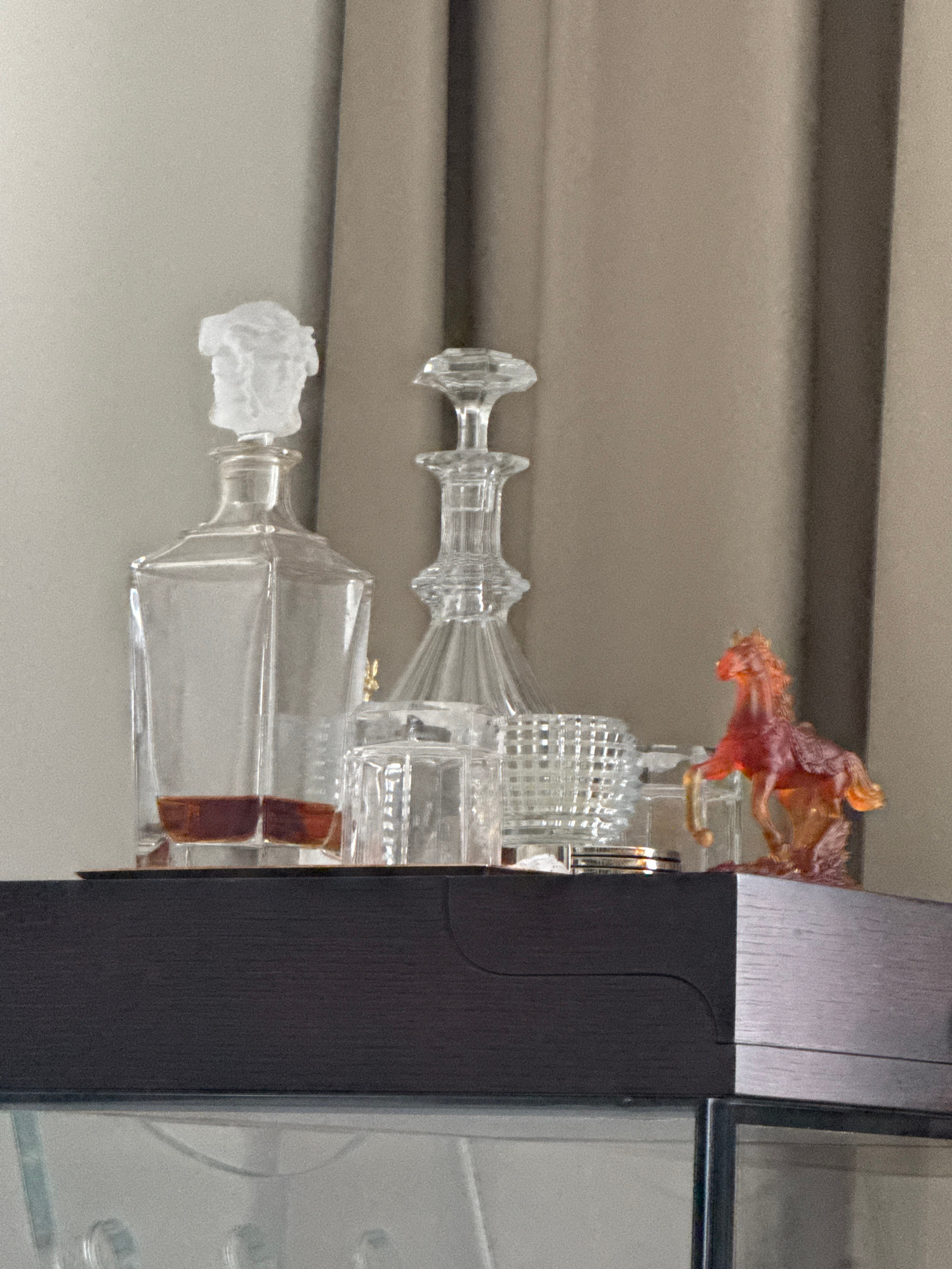 The image shows a shelf with several glass objects. On the left, there is a clear glass decanter with a frosted bust stopper, containing a small amount of amber-colored liquid at the bottom. Next to it are two clear glass tumblers. To the right of the tumblers is another clear glass decanter, this one with a more ornate stopper and a faceted body. In front of the second decanter and to the right, there is a small, translucent amber-colored glass figurine of a horse in a rearing pose. The objects are placed on a dark wooden surface, which appears to be a shelf or the top of a cabinet. Behind the objects, there are draped curtains of a neutral, beige color. The overall scene suggests a decorative display, possibly in a home bar or living area. The image does not provide any information that would allow for the determination of its location in Moscow, Russia.
