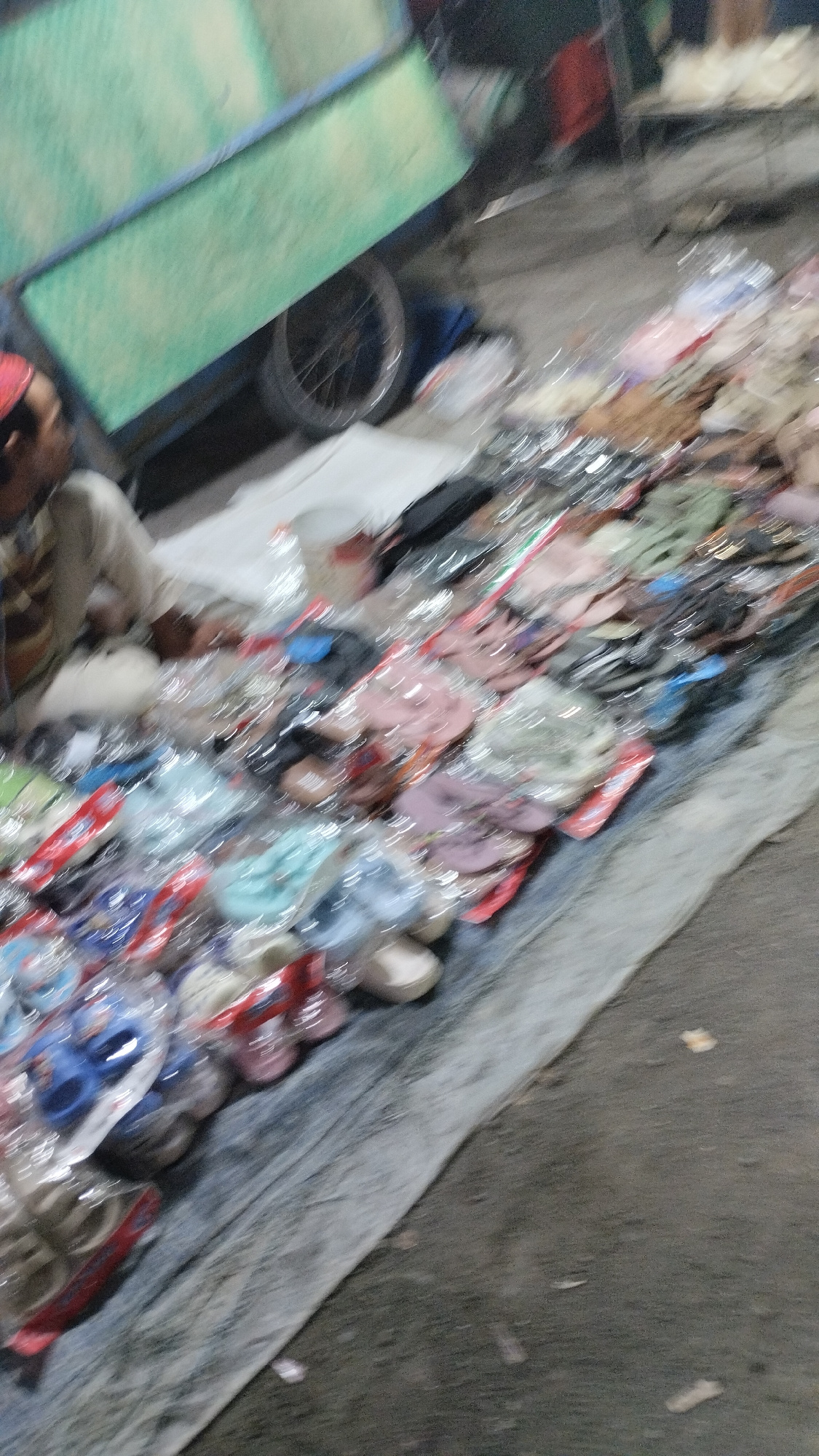 The image depicts a street vendor in Sambungrejo, Indonesia, selling footwear. A person wearing a red cap and a light-colored shirt is partially visible on the left, seemingly attending to the stall. The stall itself is a long table covered with a gray tarp, laden with rows of footwear, mostly sandals and flip-flops, displayed in clear plastic packaging. The items are arranged in a visually appealing manner, with various colors and styles visible.
Behind the stall, a green cart with a blue frame is visible, possibly used for transporting goods. The cart has a large wheel, suggesting it might be a type of pushcart or a mobile stall. The background is blurred, but it appears to be an outdoor setting, likely a street or market. The ground is rough, with visible dirt and debris, indicating an unpaved area.
The lighting suggests it could be daytime, though the image quality is low and blurred, making it difficult to determine the exact time or weather conditions. The overall impression is that of a busy marketplace, with the vendor actively engaged in selling their merchandise. The focus is on the array of footwear, showcasing the variety available to potential customers. There is no visible text in the image. The scene conveys a sense of everyday commerce and local entrepreneurship.