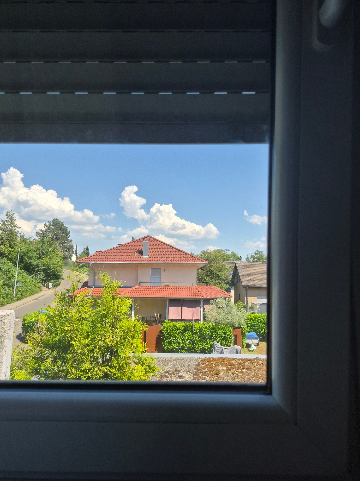 The image, captured from an interior perspective looking outwards through a window, depicts a sunny daytime scene with a clear blue sky filled with fluffy white clouds. The view is framed by the dark interior of what appears to be a room, with blinds visible on the left side of the frame.
The primary subjects are a few residential buildings with red tile roofs, surrounded by lush green trees and manicured hedges. One prominent house has light-colored walls and a balcony. A paved road winds through the scene, bordered by vegetation. In the foreground, a vibrant green tree with feathery foliage is prominently featured. On the right side of the frame, a textured surface, possibly a wall or a rooftop, is visible. The overall impression is one of a peaceful suburban or village setting on a bright, fair-weather day. No people are visible in the frame, and there is no discernible text. The time of day is clearly indicated by the bright sunlight casting shadows, and the weather is pleasant.