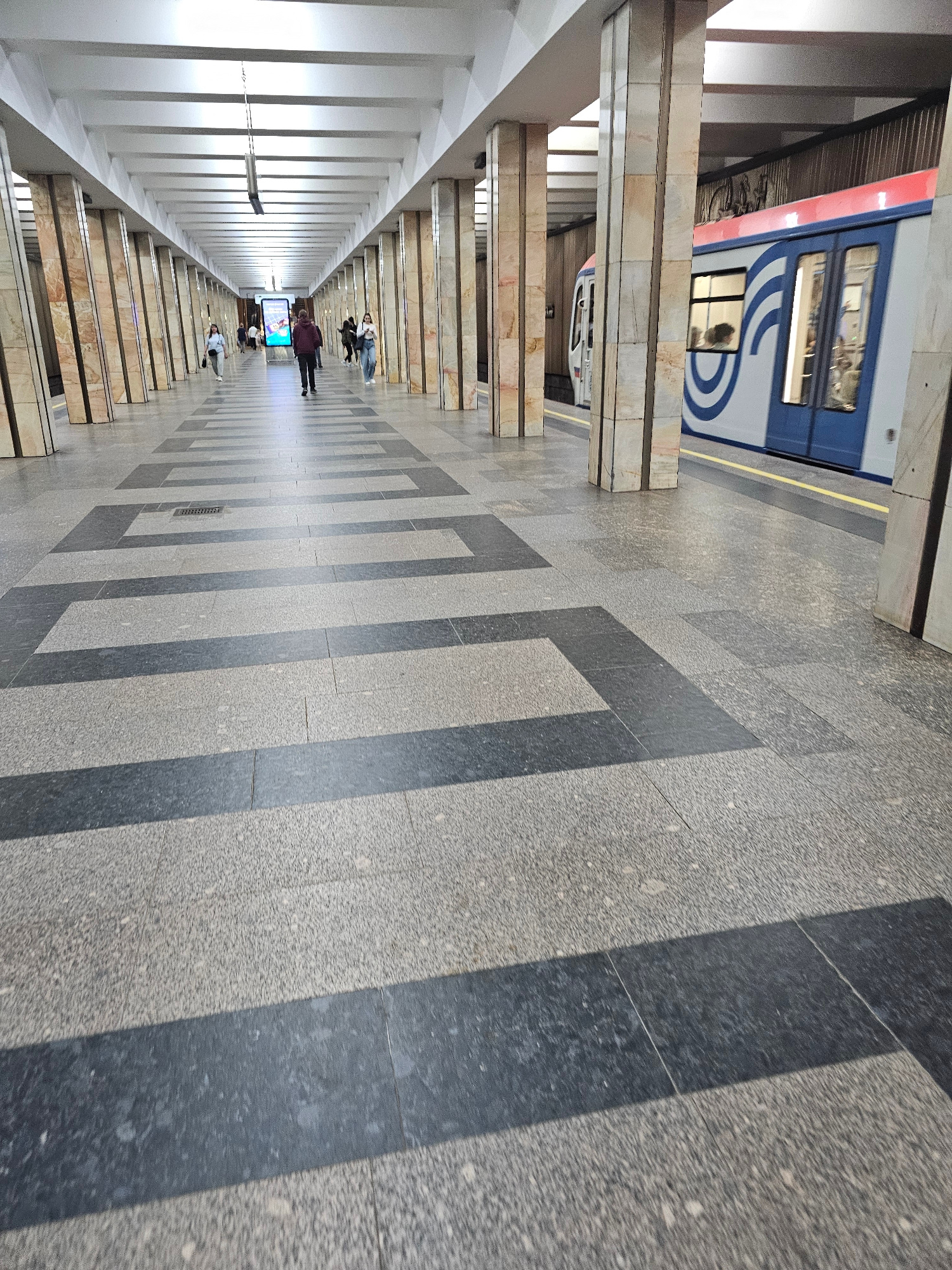This is a low-angle, vertically oriented photograph taken inside a subway station. The scene is a long, wide platform with a train visible on the left side. The platform floor is paved with speckled grey tiles interspersed with dark grey rectangular patterns. Along the right side of the platform, there are columns that support an overhead structure, creating a textured pattern of light and shadow.
Several people are visible on the platform. In the foreground and mid-ground, there are groups of individuals walking or standing, appearing to be commuters. One person in a dark jacket is walking towards the camera. Further down the platform, a group of people are gathered, with some looking at a digital screen that is illuminated.
The train on the left is modern, with blue and white detailing and red accents. Its windows are tinted, and a hint of movement is visible, suggesting it is either arriving or departing. The lighting in the station appears to be artificial, with bright lights illuminating the ceiling and the left side of the platform. The overall mood is utilitarian and busy, typical of a public transportation hub. There is no visible text in the image. The time of day and weather are not discernible from the interior setting.