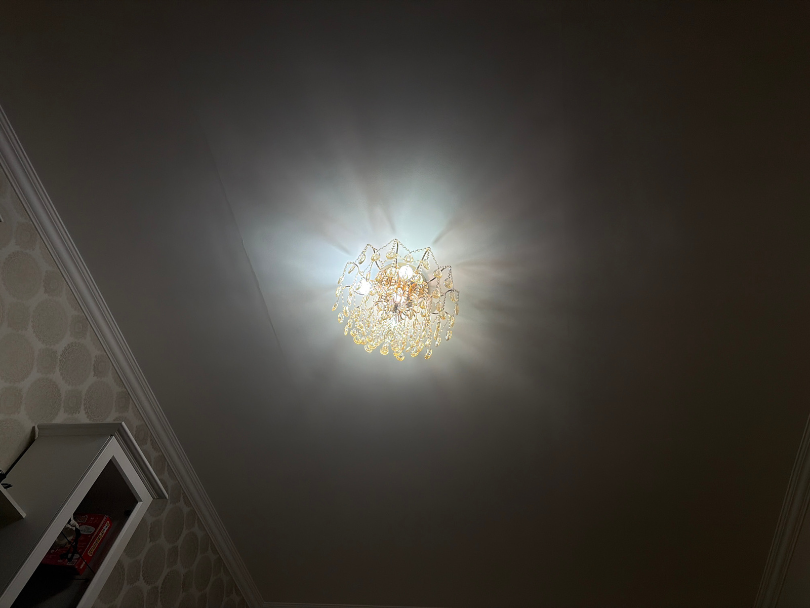 An indoor ceiling fixture, a chandelier with multiple crystalline elements, hangs illuminated against a dark ceiling. The chandelier casts a bright light, creating a halo effect. To the left, a portion of a white shelf unit is visible, containing a red box with some visible text. The walls are adorned with a patterned wallpaper. The image appears to be taken in a residential setting. There is no indication of the city of Astana, Kazakhstan.