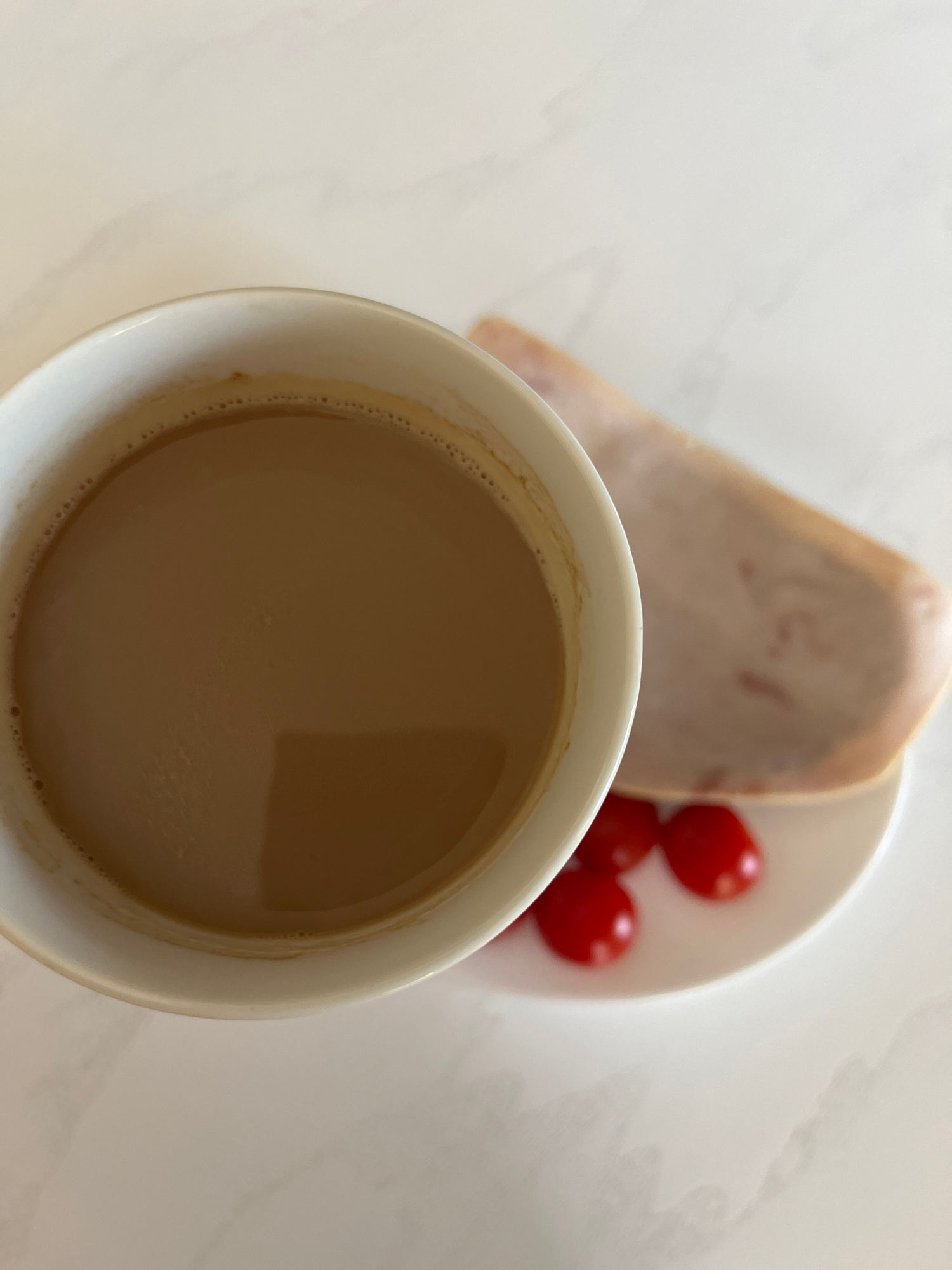 A white ceramic mug filled with coffee sits on a light-colored marble surface. To the right of the mug is a small white saucer with three red cherry tomatoes and a slice of light-colored meat, possibly ham or turkey. The image is taken from a top-down perspective, with the mug partially obscuring the saucer and its contents. There are no people visible in the image. The lighting is bright, creating soft shadows on the marble surface. The image does not provide any information about its location in Novosibirsk, Russia.