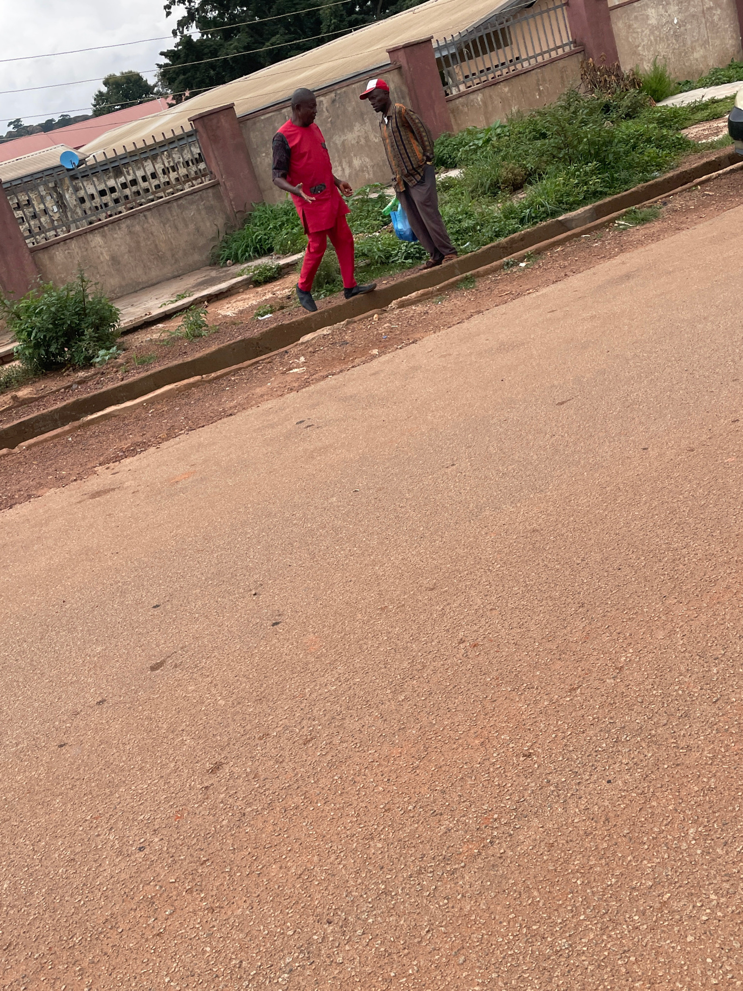 In Gura Top, Nigeria, two individuals are the focus of this media. One person, wearing a vibrant red outfit with black accents on the sleeves and a red cap, stands on a paved surface. Their posture suggests they are in mid-conversation or explaining something, with one hand raised. Beside them, another person, dressed in a patterned top and dark trousers, stands in a more relaxed pose, holding a blue object.
The scene appears to be an outdoor setting, likely a street or pathway, with a building visible on the left side. The building has a weathered concrete facade and metal bars over windows or openings. Lush green vegetation lines the edge of the paved area, suggesting a natural environment contrasting with the urban elements. A drainage channel runs alongside the pavement. The ground is covered with a reddish-brown asphalt or concrete material, indicative of a warm climate. The lighting suggests it is daytime, possibly overcast as the sunlight is diffused. The overall mood is neutral, focusing on the individuals and their surroundings. No visible text is present in the image.