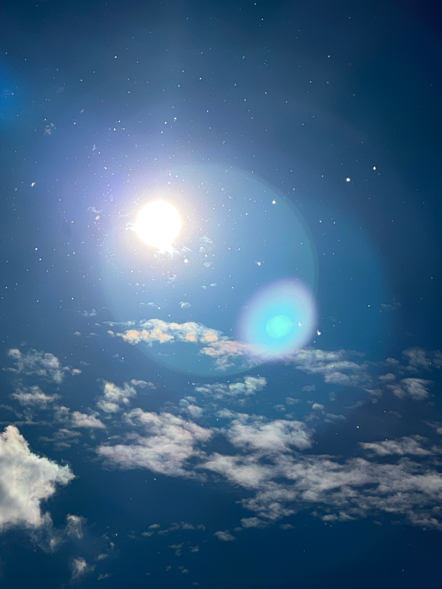 A bright, circular light source, likely the sun or moon, dominates the upper left quadrant of the image. It emits a strong lens flare, creating concentric rings of light. Scattered across the dark blue sky are numerous small, white specks, which could be dust particles, stars, or digital artifacts. In the lower half of the image, a band of scattered clouds stretches across the frame. These clouds are illuminated from above, with their edges appearing bright white and their undersides tinged with the dark blue of the sky. The clouds are wispy and irregular in shape. A faint, circular, teal-colored lens flare is visible in the middle right of the image, overlapping with the clouds. The overall impression is that of an upward-facing shot of a sky with dramatic lighting and atmospheric elements.