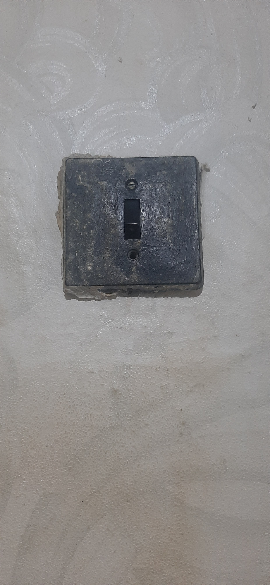 A close-up, eye-level shot shows a weathered, black, square electrical light switch mounted on a pale, textured wall in Yessentukskaya, Russia. The switch has a rectangular button in the center, with a visible line dividing it into two smaller sections, indicating it likely controls two separate circuits or lights. The switch plate is visibly dirty and worn, with chipped edges and a rough texture, suggesting age and considerable use. Two circular indentations, likely for screws, are visible on either side of the switch button, further highlighting its utilitarian design. The surrounding wall is a light, off-white color with a faint, swirling pattern etched into its surface, possibly from past repairs or decoration. The texture of the wall is slightly bumpy and uneven, with small, circular indentations scattered across it, creating a mottled appearance. The lighting in the image is even and appears to be artificial, with no strong shadows or highlights, suggesting an indoor setting, likely in a building. The scene is simple and functional, focusing entirely on the details of the aged electrical switch and the wall it is mounted on, devoid of any other objects or human presence. There is no visible text in the image.