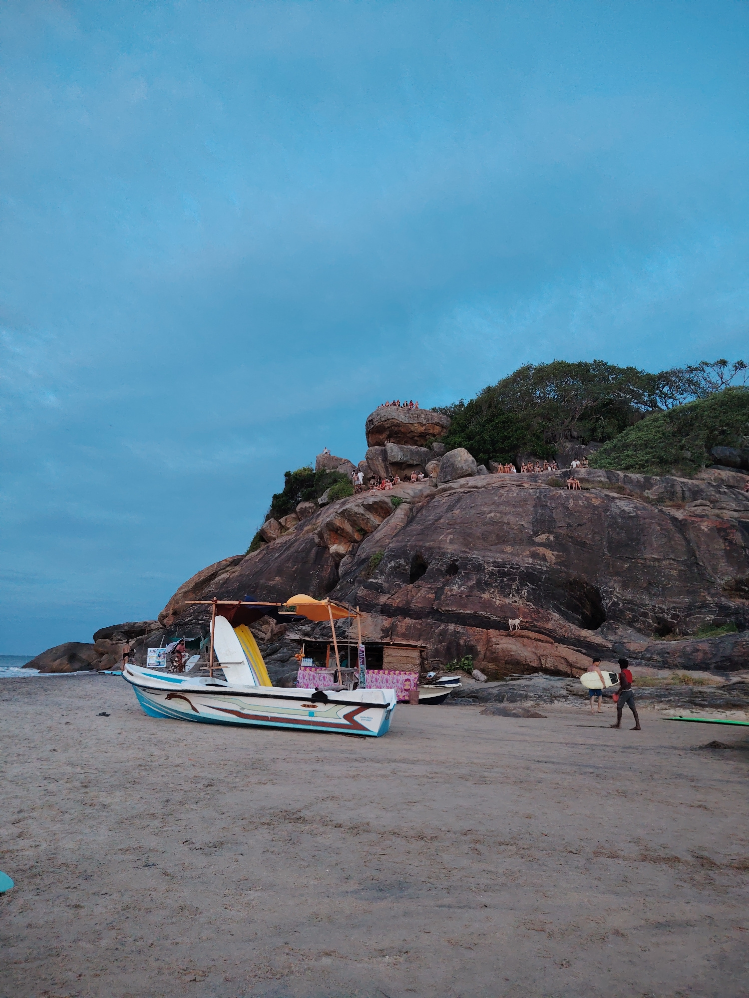This is a medium shot of a beach scene in Shastrawela, Sri Lanka, at dusk. A sandy beach stretches across the foreground, with the ocean visible to the left. A white and blue boat with a covered canopy is positioned on the sand, laden with yellow and white surfboards. Behind the boat, a stall or small structure is set up. To the right, two young men are standing on the sand, one of whom is carrying a surfboard. Further back, a large, rocky cliff rises steeply, topped with lush green foliage. Numerous people are visible gathered on various ledges of the cliff, appearing to be enjoying the view. A small, light-colored dog is walking on the rocky face of the cliff. The sky above is a deep blue, transitioning to a lighter shade near the horizon, indicating the time of day. The overall atmosphere is one of a relaxed, coastal community gathering at sunset.