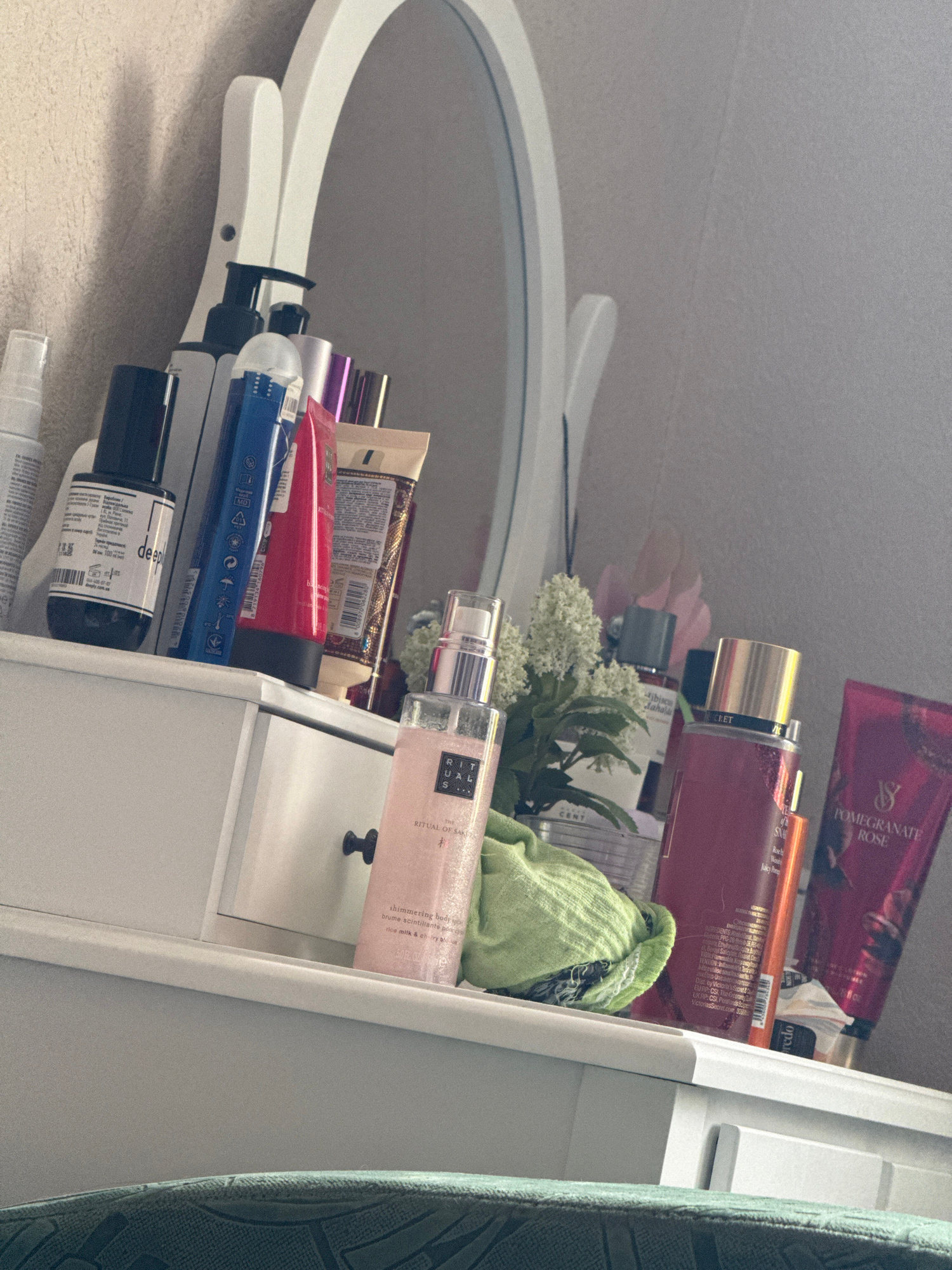 An indoor, eye-level medium shot captures a vanity dresser with various beauty products arranged on its surface. The dresser is white and has a single drawer with a black handle. The products include several bottles and tubes of lotions, perfumes, and other cosmetics. Among them are recognizable brands like Victoria's Secret and Rituals. A white oval mirror with a decorative frame is positioned behind the dresser. The background is a textured light-colored wall. The lighting appears to be natural and somewhat dim.