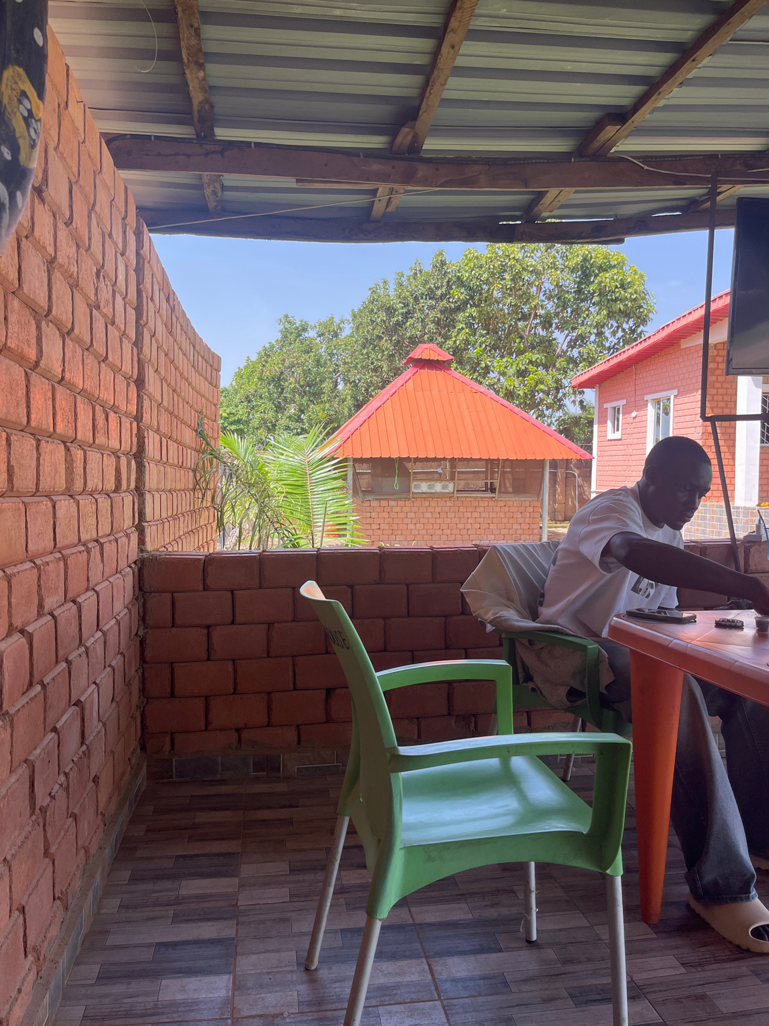 This image shows a man sitting at an orange plastic table under a corrugated metal roof supported by wooden beams. He is wearing a white t-shirt and dark pants, and is looking down at the table. A green plastic chair is in the foreground. The scene appears to be an outdoor seating area, possibly a restaurant or cafe, with red brick walls on the left. In the background, there are trees, a small building with an orange roof, and another building that is partially visible. The overall impression is a casual, outdoor setting in what appears to be a warm climate.