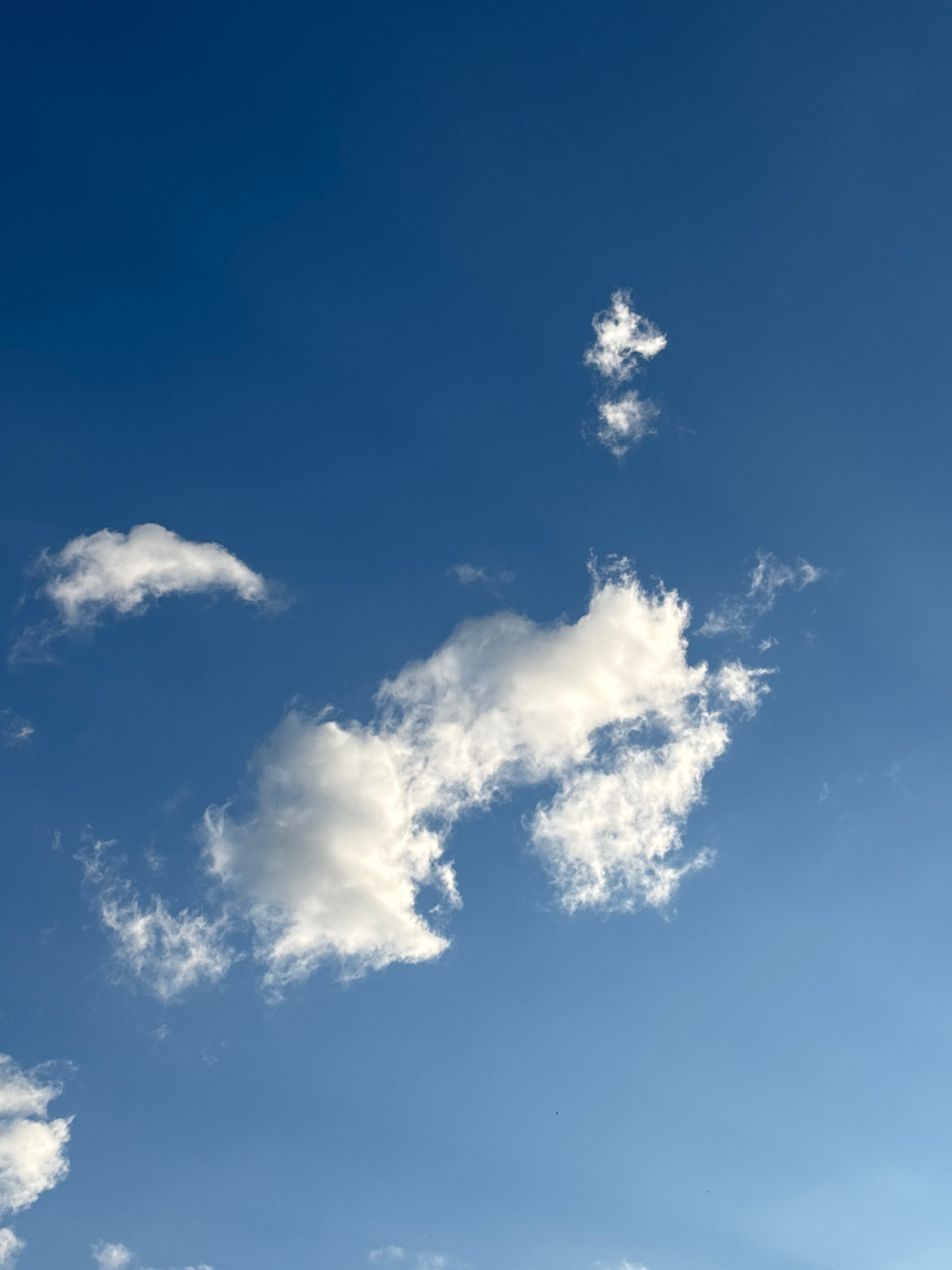 A vertical, medium shot captures a clear blue sky dotted with white, wispy clouds. The clouds are scattered across the frame, with a larger cluster towards the center and smaller formations towards the top and bottom. The lighting suggests it is daytime, with sunlight illuminating the clouds from above. The scene is devoid of any human presence or man-made objects, presenting a natural, unobstructed view of the sky.