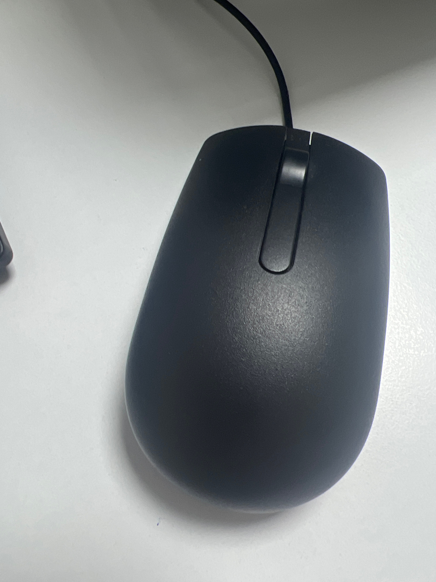 The image shows a black computer mouse on a white surface, likely a desk. The mouse is oriented with its scroll wheel towards the top of the frame. A black cable extends from the top of the mouse and disappears out of the frame. The surface appears to be textured. No people or city elements are visible in the image.