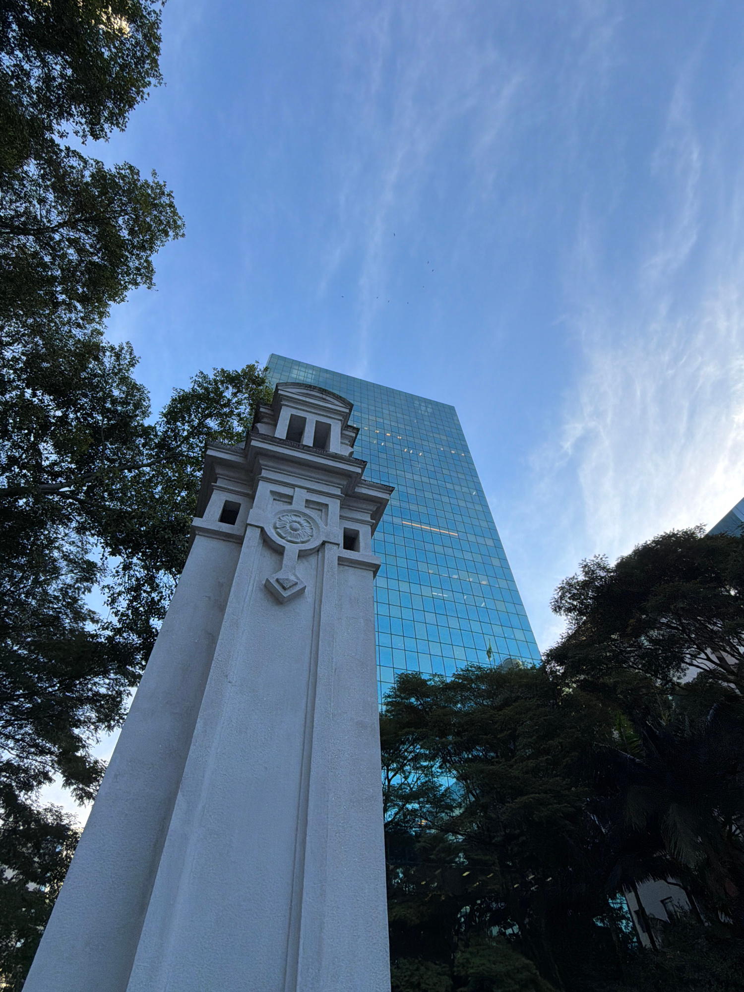 An upward shot from a low angle features a weathered, light-colored stone monument in the foreground, its top section adorned with decorative carvings, including a circular motif. Behind the monument, a tall, modern glass-facade skyscraper dominates the background, its blue-tinted windows reflecting the sky and surrounding trees. Lush green trees partially obscure the skyscraper on the left and right sides of the image. The sky above is a clear blue with wispy white clouds, and a few small birds are visible as specks. The scene is set outdoors, possibly in an urban park or public space within São Paulo, Brazil, given the architectural juxtaposition of historical and contemporary elements.