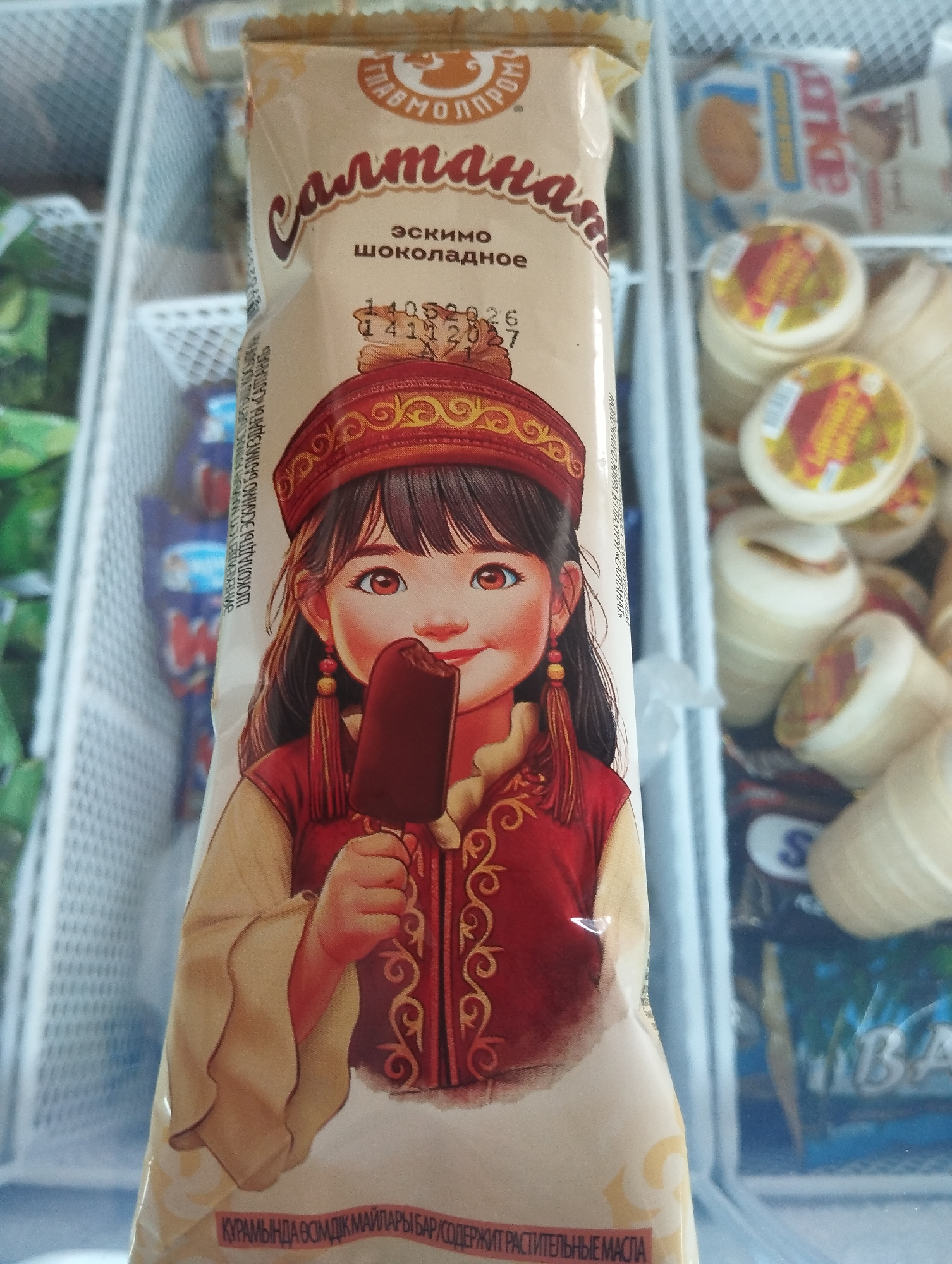 A close-up, slightly low-angle, indoor shot shows a popsicle wrapper featuring a cartoon illustration of a young girl in traditional attire holding a chocolate-covered ice cream bar. The girl has dark hair, large brown eyes, and wears a red hat with gold trim and a red vest with gold embroidery over a cream-colored garment. She is holding a dark brown popsicle up to her mouth. The wrapper is light brown and features text in Cyrillic script, including "Салма", "Эскимо Шоколадное", and a date "14 05 2026". To the right and in the background, various other frozen treats are visible in a freezer case. The setting appears to be a retail environment in the city of Kojomkul, Kyrgyzstan, likely a grocery store or convenience store.