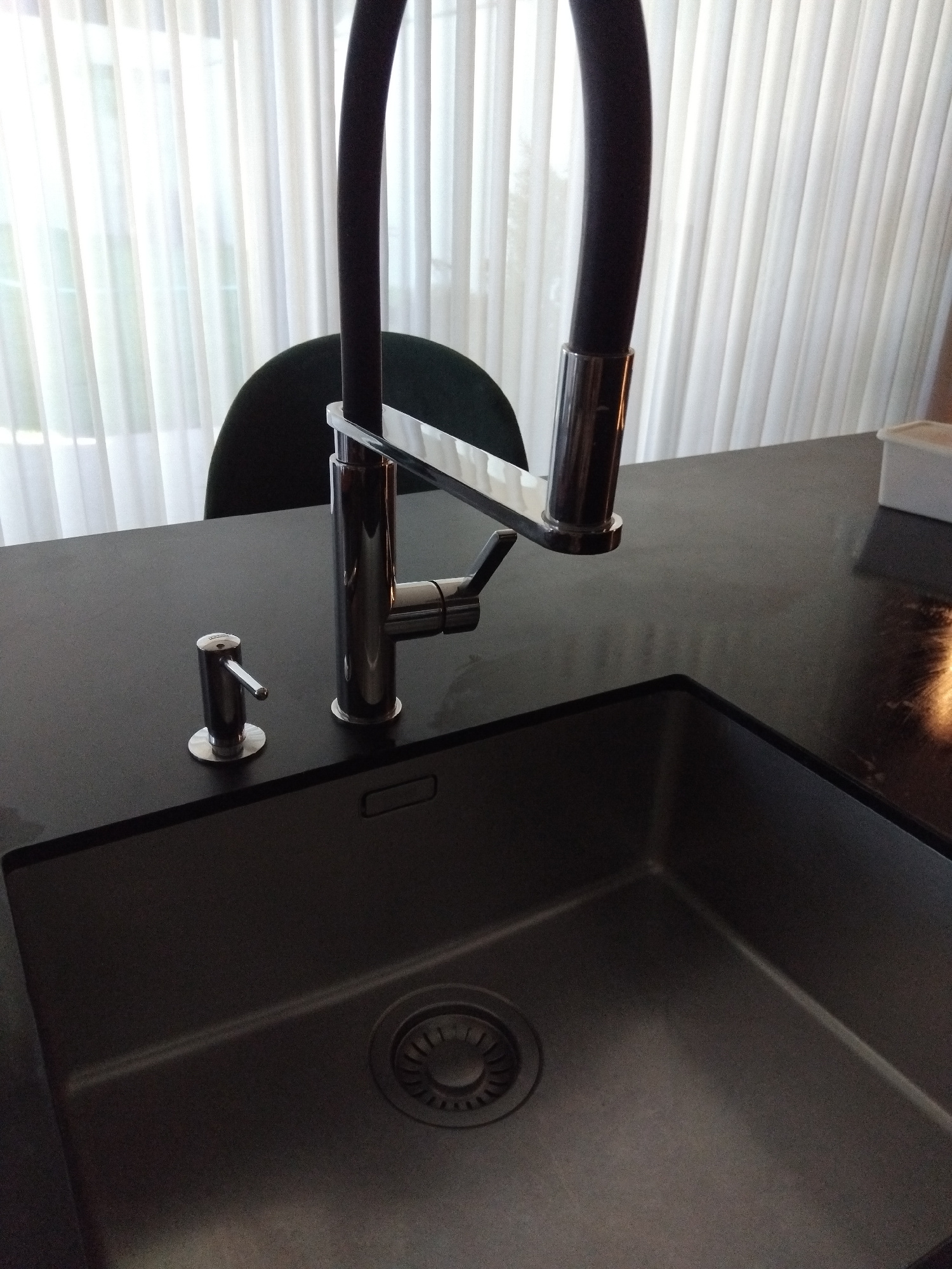 A high-angle, medium shot captures a modern kitchen sink area with a sleek, dark countertop and a metallic faucet. The faucet is tall and features a flexible black hose. To the left of the faucet, a chrome soap dispenser with a pump mechanism is visible. The sink itself is deep and metallic, with a drain at the bottom. In the background, sheer white curtains hang, diffusing natural light and creating a soft backdrop. The overall scene suggests a clean, contemporary kitchen environment.