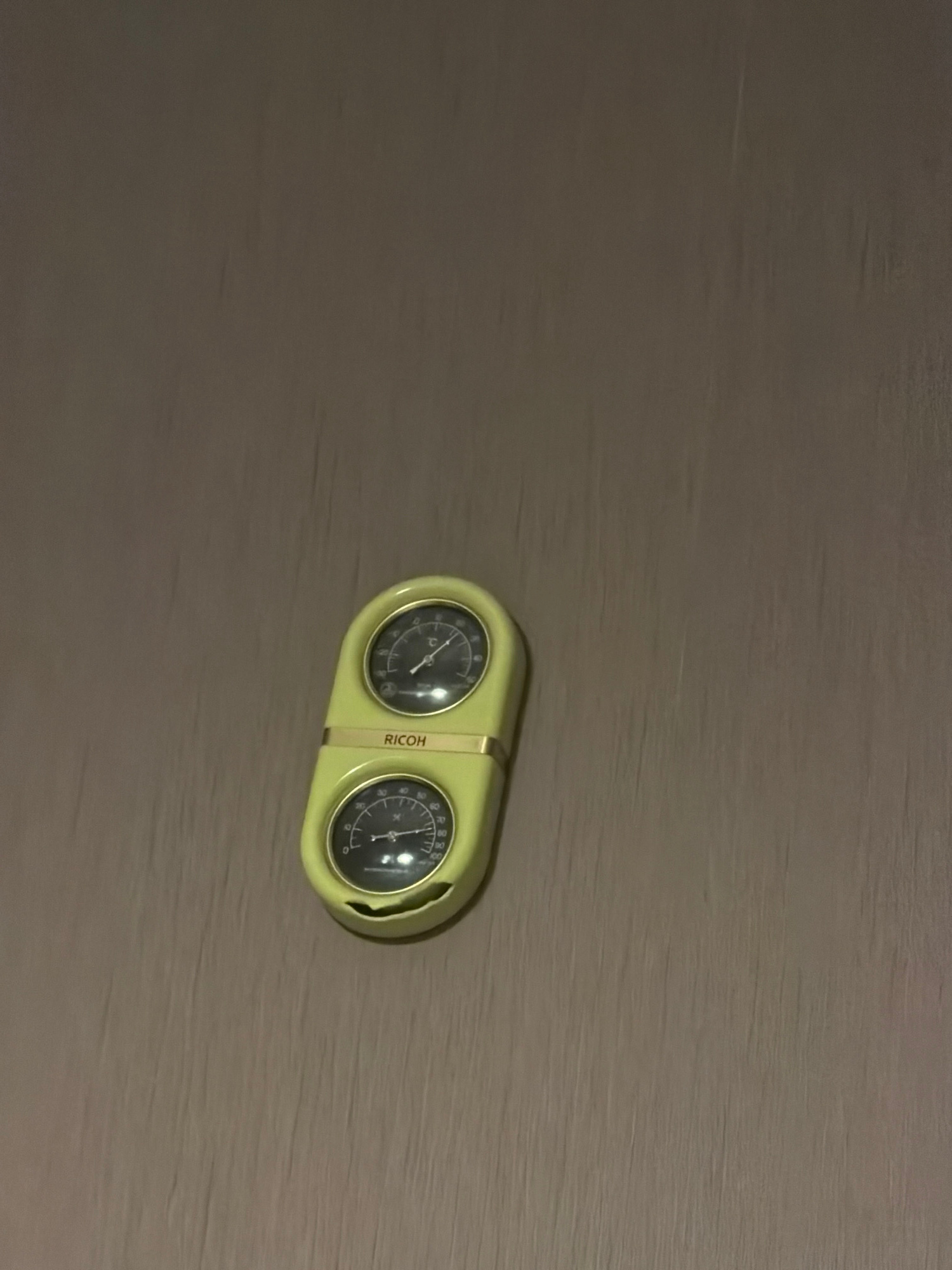 A yellow, oval-shaped object with two circular dials is mounted on a textured, brown wall. The object has a gold trim separating the two dials. The upper dial has a needle pointing to approximately 20 degrees Celsius. The lower dial has a needle pointing to approximately 60% humidity. The word "RICOH" is printed in gold lettering between the two dials. The wall appears to be made of a material with vertical striations, possibly wallpaper or painted drywall. The lighting is dim, casting subtle shadows. There are no people or other objects visible in the frame.