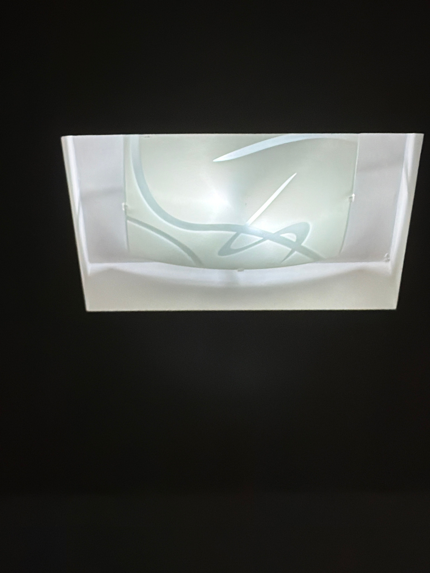A flush-mount ceiling light fixture is visible against a dark background. The fixture has a rectangular frame and a frosted glass cover with a decorative pattern of swirling lines in a lighter shade of white. The light is on, illuminating the glass. The scene is indoors and appears to be in a residential or commercial space. There are no people or other objects clearly discernible in the image, nor is there any indication of a specific location within Plovdiv, Bulgaria.