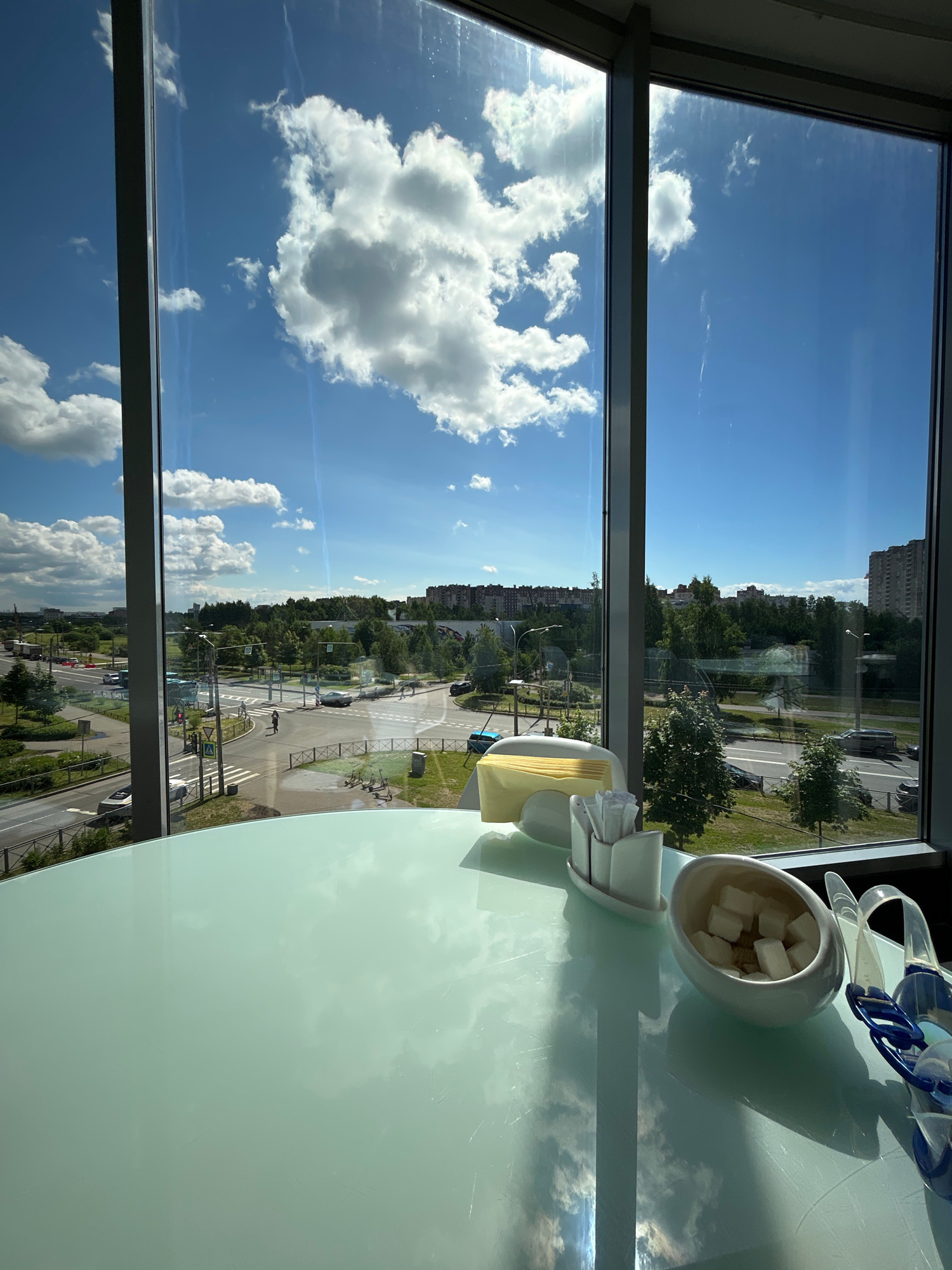 The image shows a view through a large window from an elevated position, looking out over a cityscape. In the foreground, on a light-colored, reflective surface, are a sugar bowl filled with sugar cubes, a dispenser for sugar packets or napkins, and a pair of swimming goggles. The window looks out onto a street scene with traffic, trees, and apartment buildings in the distance. The sky is blue with fluffy white clouds. The specific location in Saint Petersburg, Russia cannot be determined from the image alone.