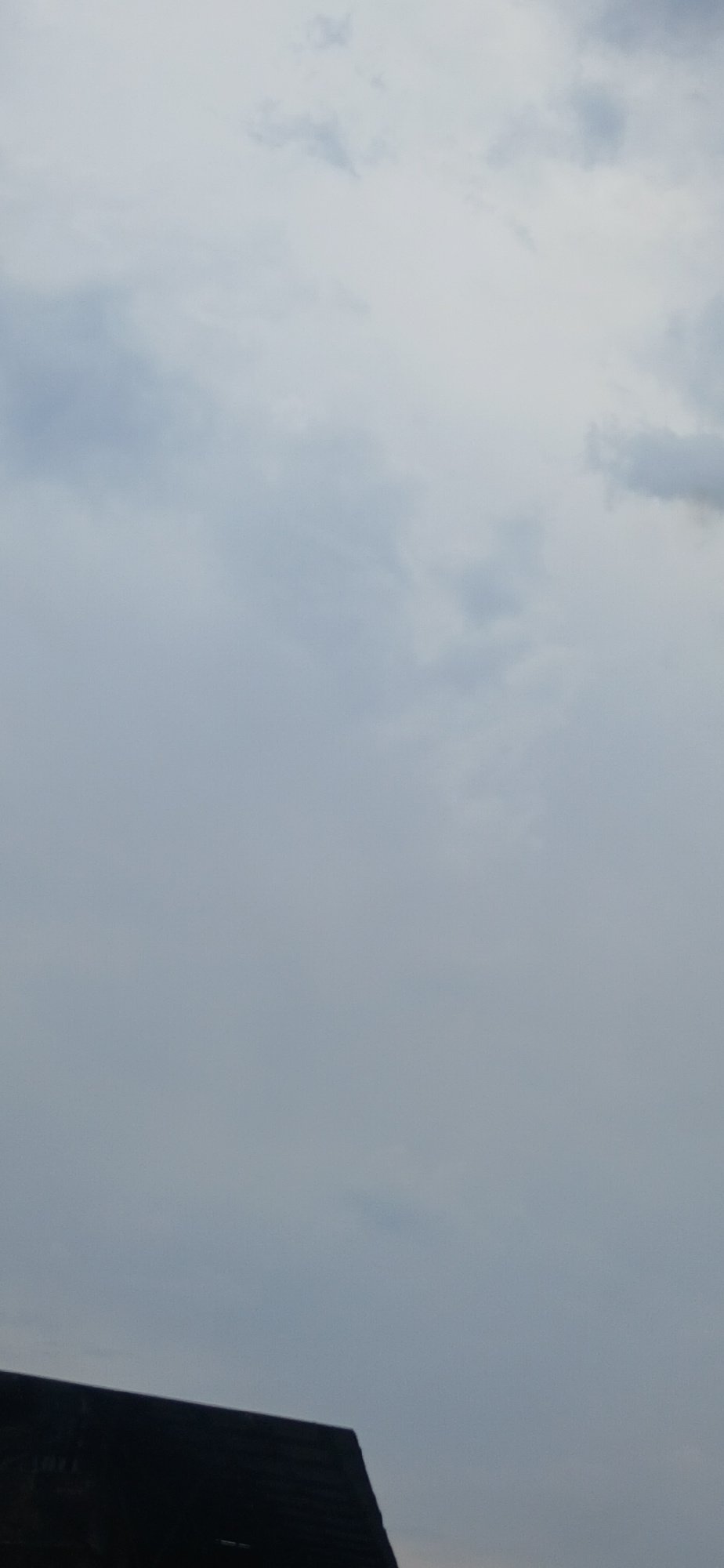 The image shows a dark, angled roofline in the lower left corner, partially obscuring a hazy, overcast sky. The sky is filled with diffused light, suggesting it is daytime but possibly cloudy or foggy. The clouds are varied in shade, with some appearing lighter and more wispy, while others are darker and more solid. No people or specific landmarks are visible. The image appears to be a photograph taken from ground level, looking upwards towards the sky and capturing a portion of a building's roof. The overall impression is one of muted light and a possibly unsettled weather condition.