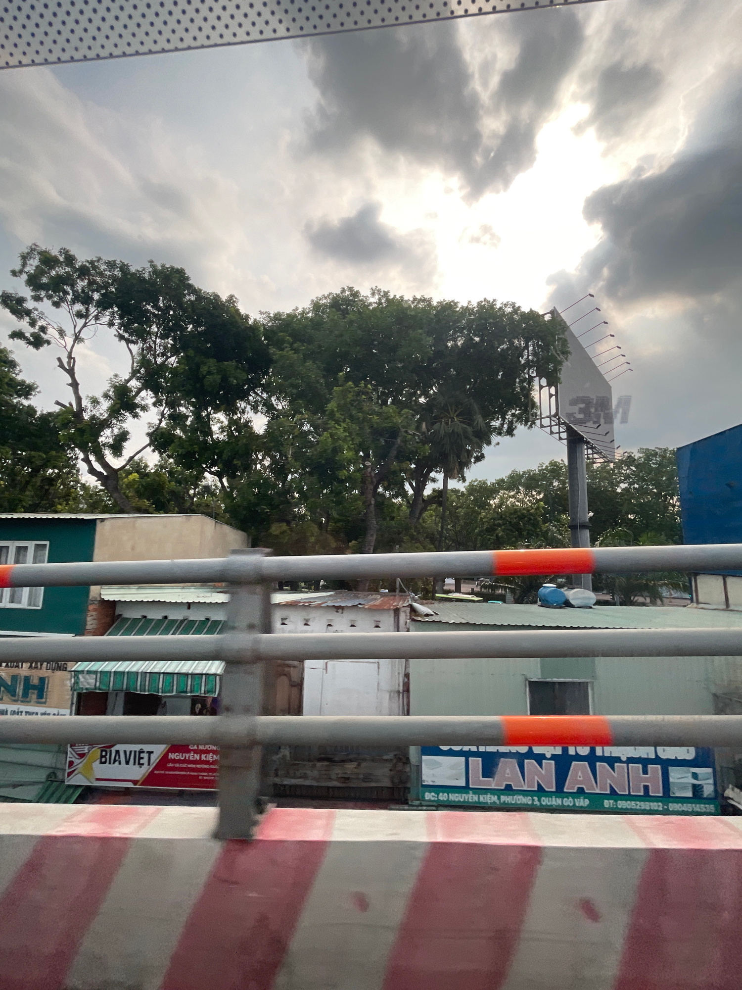 This is a vertical, eye-level, medium shot capturing a view from a moving vehicle, likely a car or bus, looking towards a street scene in Thủ Đức, Vietnam. The foreground is dominated by a grey metal railing with a red and white striped concrete barrier below it. Behind the railing, various structures and elements of the urban environment are visible.
On the left, a building with a teal facade and white-framed windows is partially visible. To its right, a beige building with a flat roof sits behind it. Further back, there are several other buildings with corrugated metal roofs and various signage. One prominent sign displays "BIA VIET NGUYEN KIEM" in red and yellow lettering. To the right of this, a large blue billboard with the text "LAN ANH" in large, stylized white letters on a blue background is clearly visible. Below this text, an address and phone numbers are listed: "DC: 40 NGUYEN KIEM, PHUONG 3, QUAN GO VAP" and "DT: 0905258102 - 0904514535".
Behind the buildings, a dense canopy of green trees forms a backdrop. Above the trees, the sky is filled with dramatic, dark clouds with patches of bright sunlight breaking through, suggesting either dawn, dusk, or an impending storm. A large billboard structure, presumably for advertising, stands tall behind the trees, with the faded "3M" logo visible on its angled surface. The overall impression is of a bustling, somewhat gritty urban landscape under a dynamic sky.