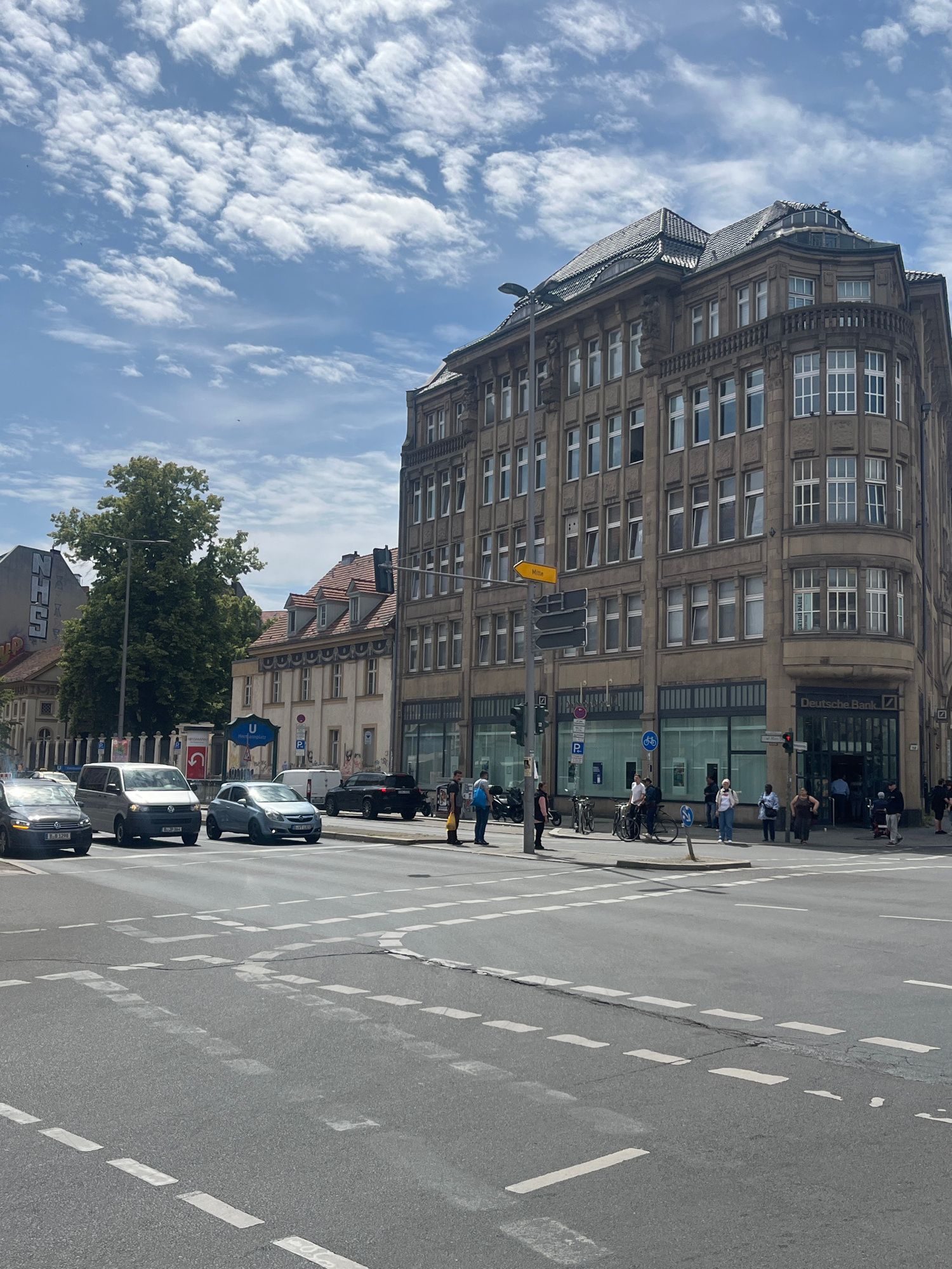 The image depicts a street scene in Berlin, Germany, on a cloudy day. In the foreground, a four-way intersection is visible with white dashed lines painted on the asphalt to delineate traffic lanes and crosswalks. Several vehicles, including cars and a van, are present on the road, some stopped at traffic lights.
The dominant feature of the scene is a large, multi-story building with a distinctive architectural style, featuring a rounded corner and numerous windows. The building appears to house a Deutsche Bank branch, indicated by signage. Adjacent to this building, other structures with pitched roofs and dormer windows are also visible, contributing to the urban landscape.
Several individuals are seen on the sidewalks and near the intersection. Some are walking, while others appear to be waiting. Bicycles and motorcycles are also parked along the street. The environment suggests a typical urban setting with commercial activity and pedestrian traffic. The overall impression is of a bustling city intersection under a partly cloudy sky.