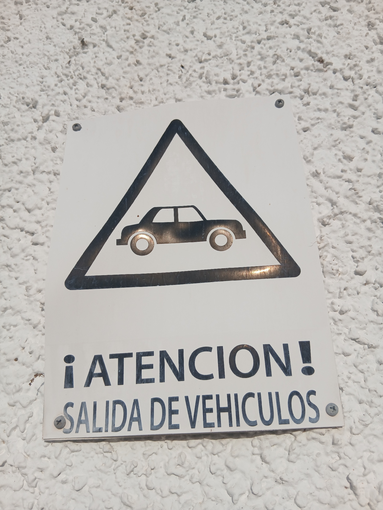 The image shows a white rectangular sign with a black border and a black outline of a car inside a black triangle. Below the triangle, the sign displays the text "¡ATENCION!" in large black letters and "SALIDA DE VEHICULOS" in slightly smaller black letters. The sign is attached to a textured white wall with screws. The lighting suggests it is daytime. The sign is likely located in Navaluenga, Spain, as indicated by the context of the request, warning of a vehicle exit.